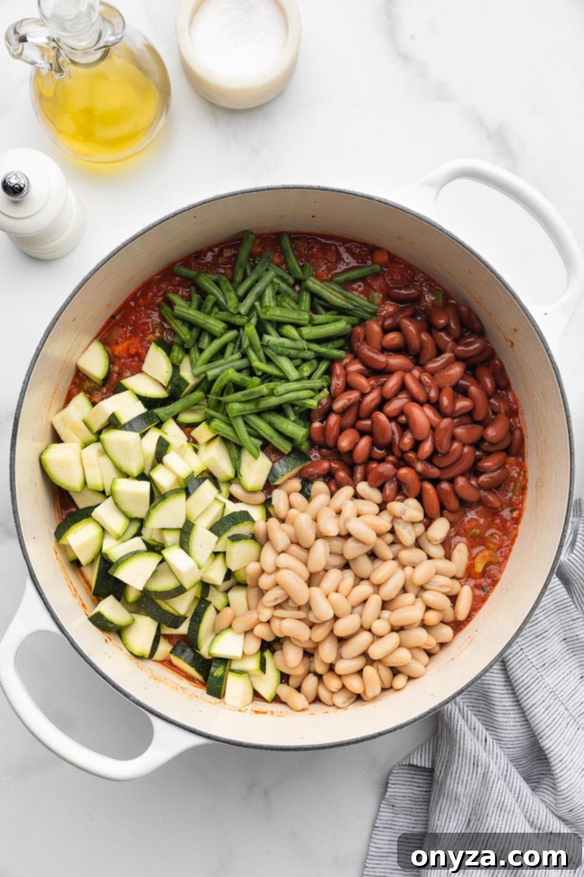 Various piles of zucchini, green beans, kidney beans, and cannellini beans added to a pot of uncooked Minestrone Soup, ready to be stirred.
