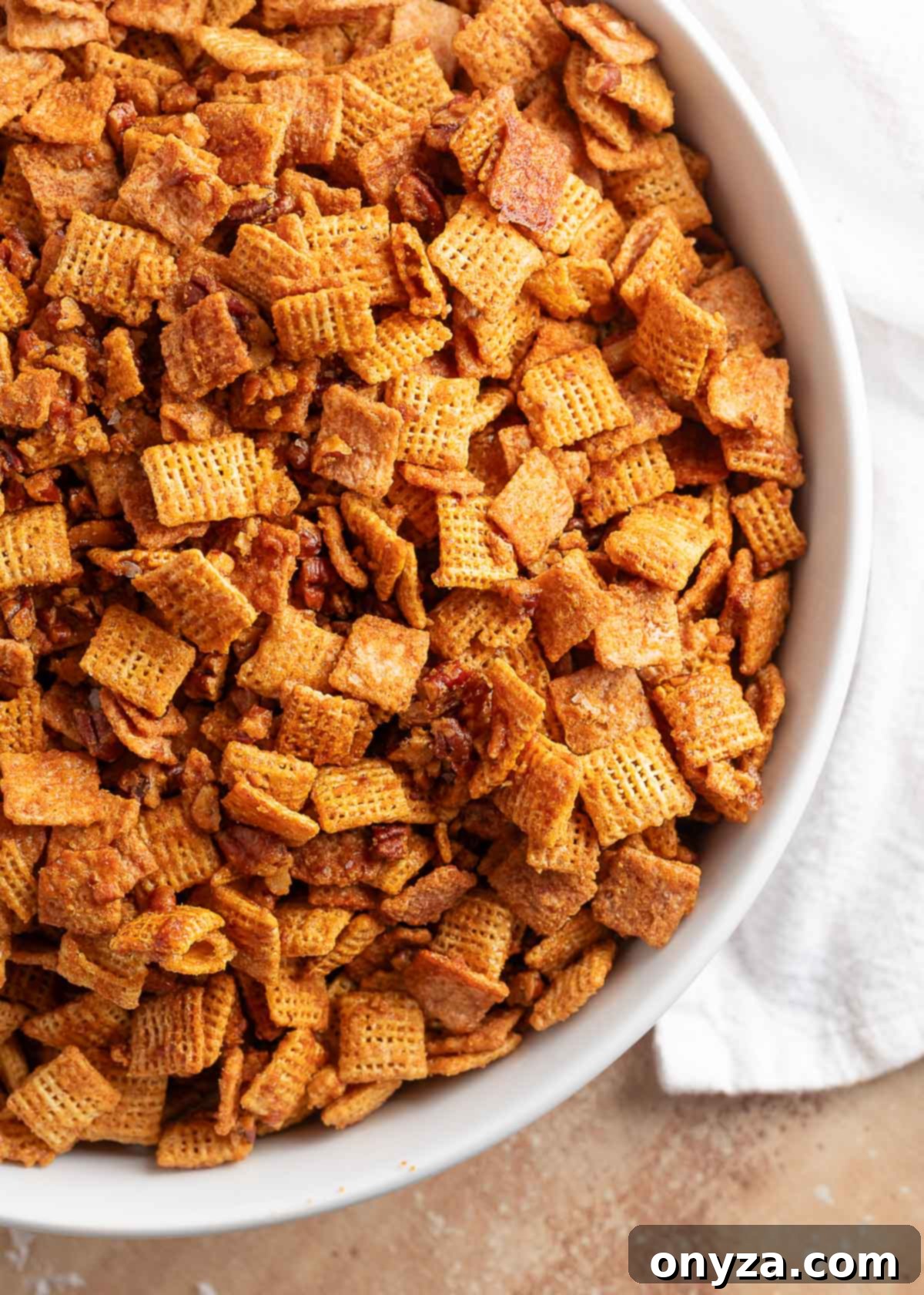 A generous white bowl filled with glistening Cinnamon Praline Crunch Snack Mix, featuring crispy cereal squares and whole pecans coated in a rich, golden glaze, ready to be enjoyed.