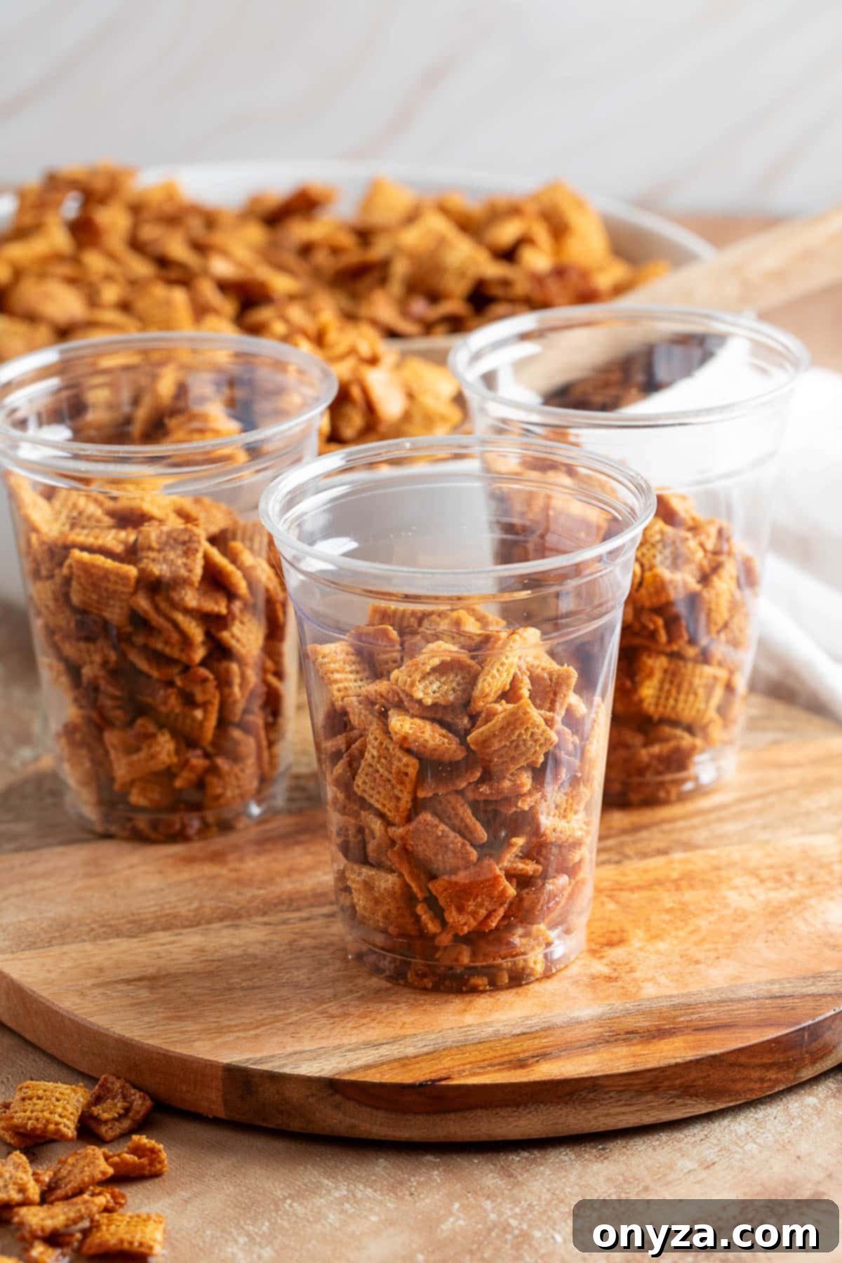 Individual clear plastic cups filled with golden cinnamon praline crunch snack mix, arranged neatly on a rustic wooden board, ready for guests to easily grab and enjoy.