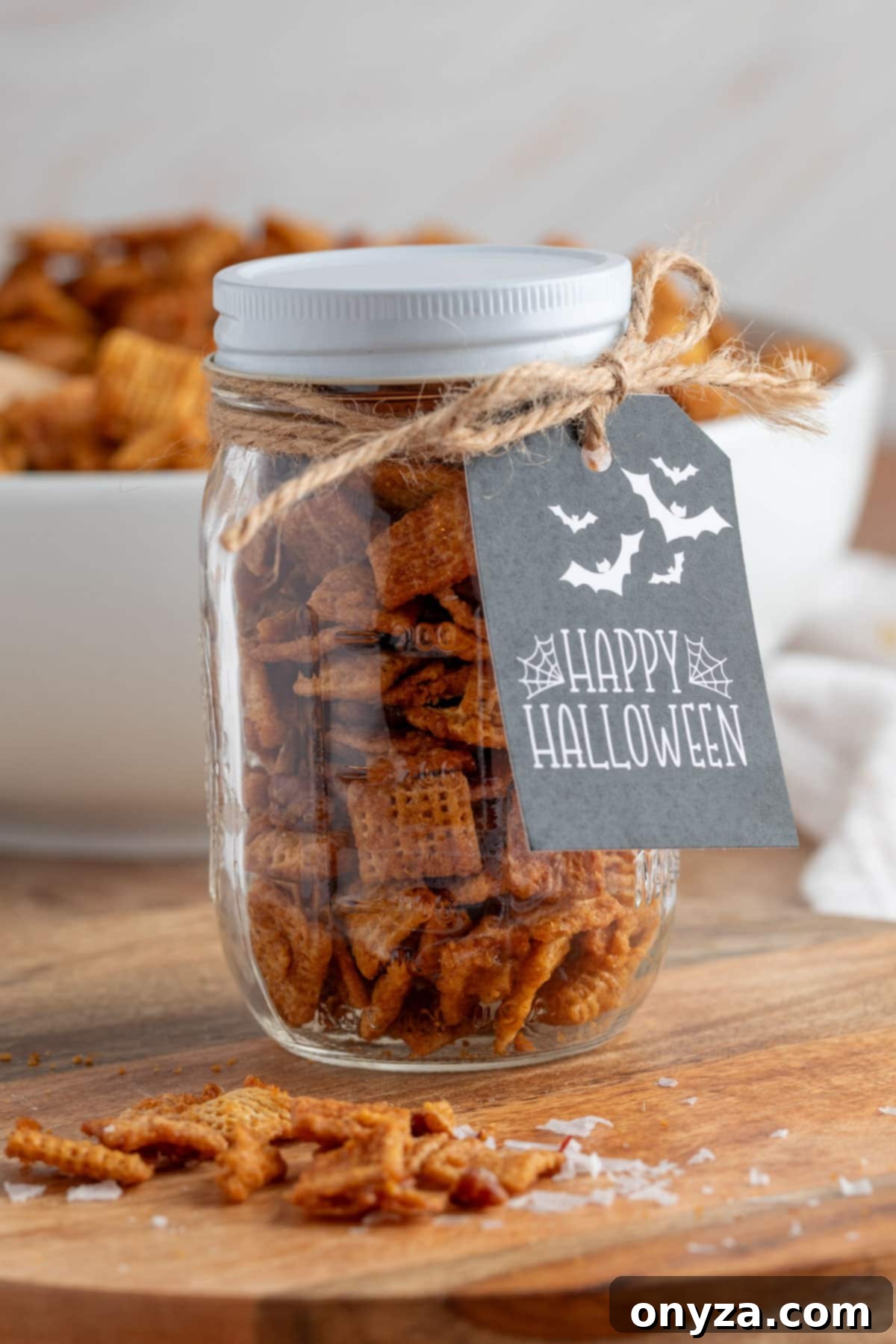 A charming glass jar, brimming with rich cinnamon praline snack mix, elegantly tied with natural twine and accented with a striking black “Happy Halloween” gift tag adorned with playful bats and delicate spiderwebs. A soft-focus bowl of the same tempting snack mix gently blurs in the background, creating a festive autumnal scene.