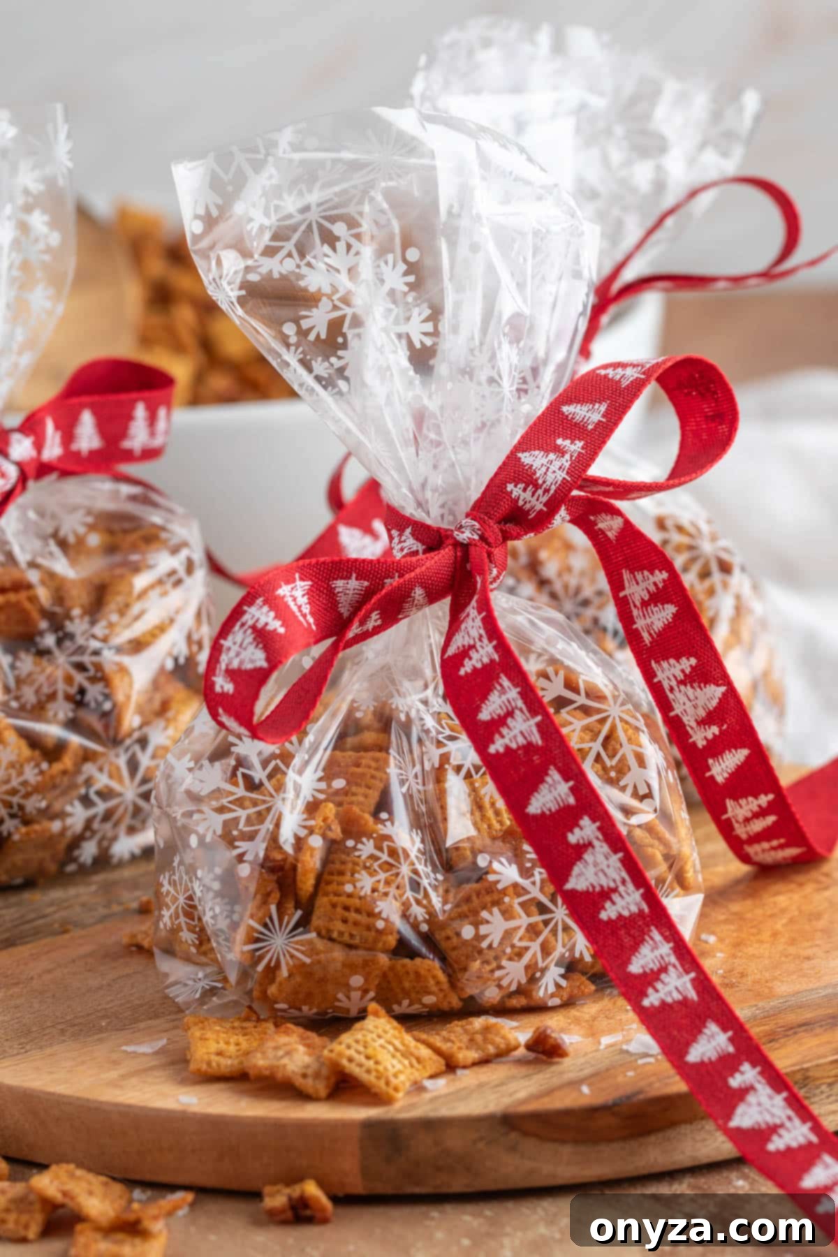 A festive display of clear cellophane gift bags, each generously filled with homemade cinnamon praline crunch snack mix, beautifully tied with vibrant red ribbon featuring delicate white Christmas trees. The bags are gracefully arranged on a rustic wooden board, with scattered snack pieces adding to the charming holiday ambiance.