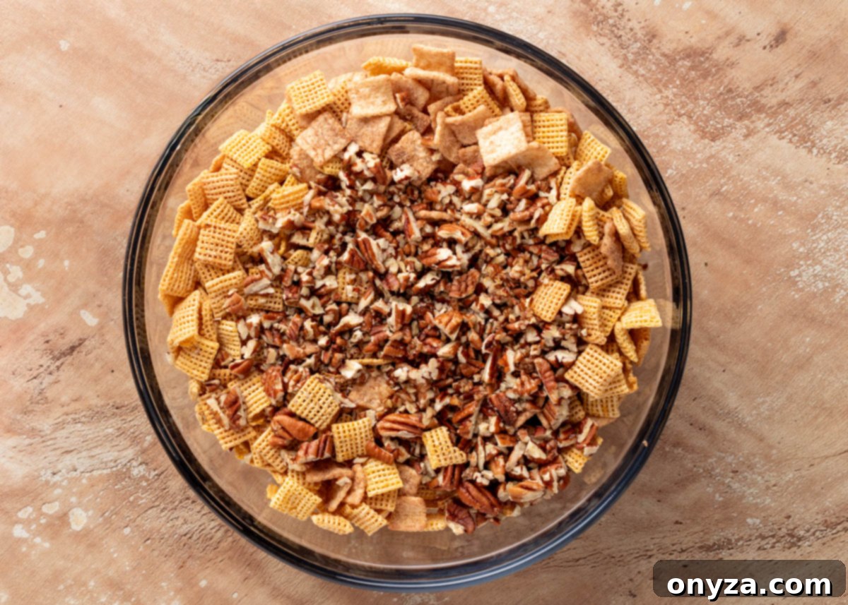 A top-down view of a clear glass mixing bowl brimming with an appetizing blend of golden corn cereal squares, crunchy cinnamon toast cereal pieces, and scattered chopped pecans, all resting on a textured tan surface, perfectly prepped and ready for the next step in making a delectable snack mix.