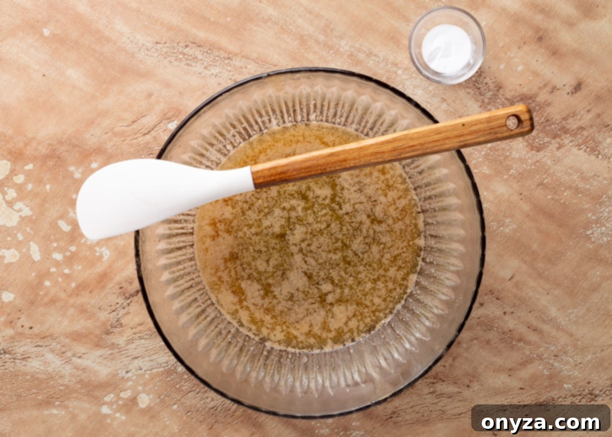 A vibrant close-up of light brown sugar, perfectly melted unsalted butter, and glistening light corn syrup swirling together in a clear glass bowl, with a white rubber spatula resting within, ready to complete the smooth praline base.