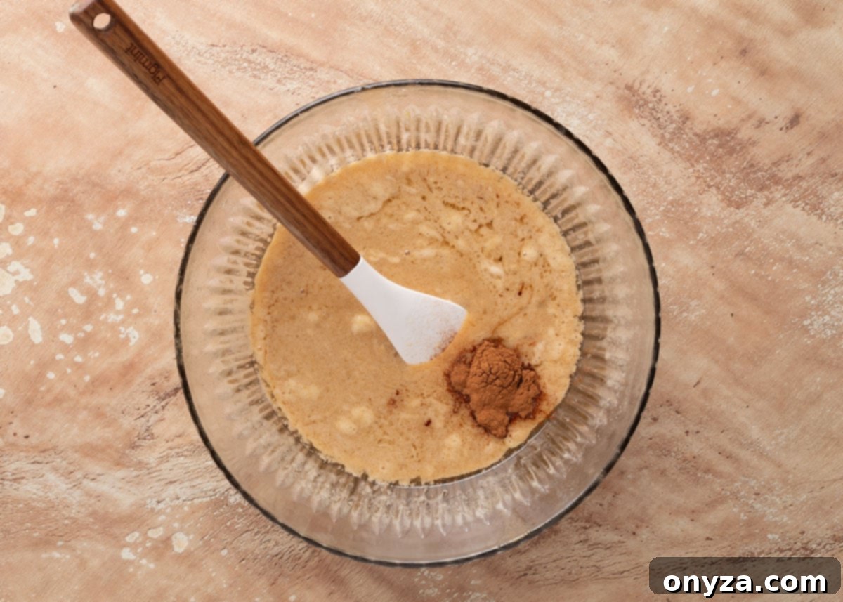 An inviting overhead view of a clear glass mixing bowl, where a bubbling butter and sugar mixture is being infused with a neat mound of ground cinnamon, ready to be incorporated by a pristine white spatula with a natural wooden handle, creating the fragrant praline glaze.