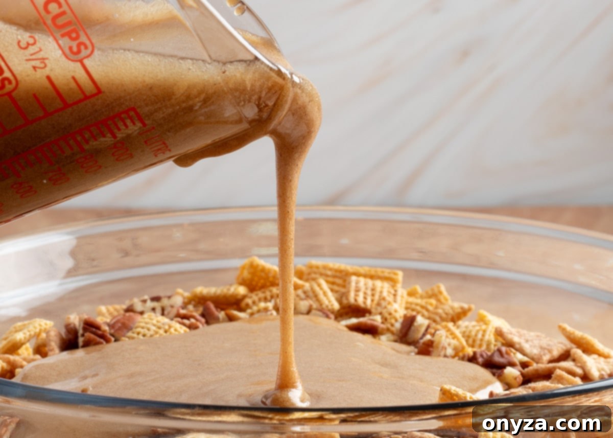 A captivating close-up of the cinnamon praline glaze gracefully cascading from a clear glass measuring cup, generously coating a luscious blend of Chex cereal and chopped pecans, all destined to become a perfectly sweet and crunchy snack mix.