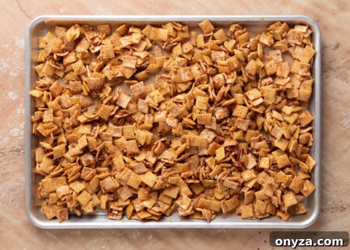 A pristine baking sheet, lined with parchment paper, holds an evenly spread layer of Cinnamon Praline Crunch Snack Mix, each cereal piece and pecan glistening with a rich caramelized coating, perfectly poised and ready for its transformative bake.