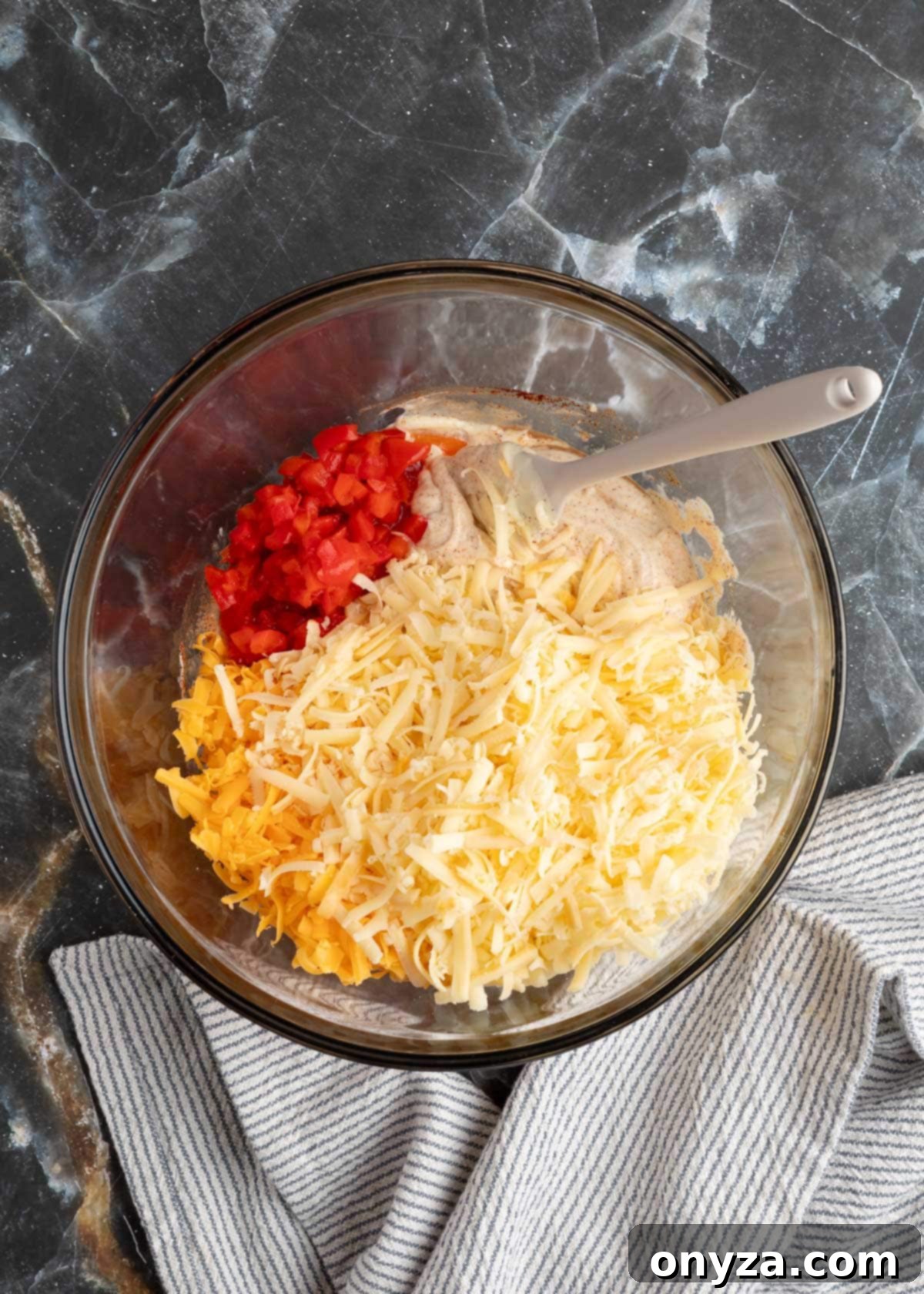 Shredded cheeses and drained pimentos being folded into the seasoned mayonnaise base in a glass bowl.