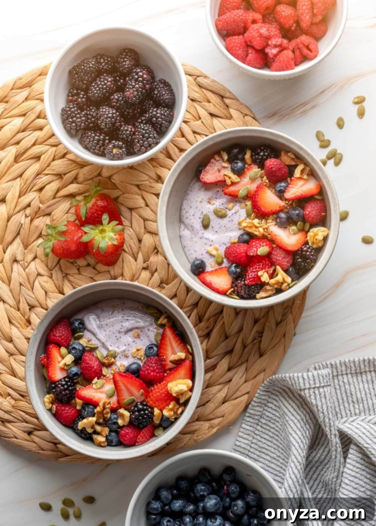 Blueberry Protein Smoothie Bowls 6 An overhead shot featuring two elegantly arranged blueberry cottage cheese smoothie bowls, generously topped with an assortment of fresh fruits and crunchy granola, resting on a woven rattan mat alongside small bowls of vibrant fresh berries.