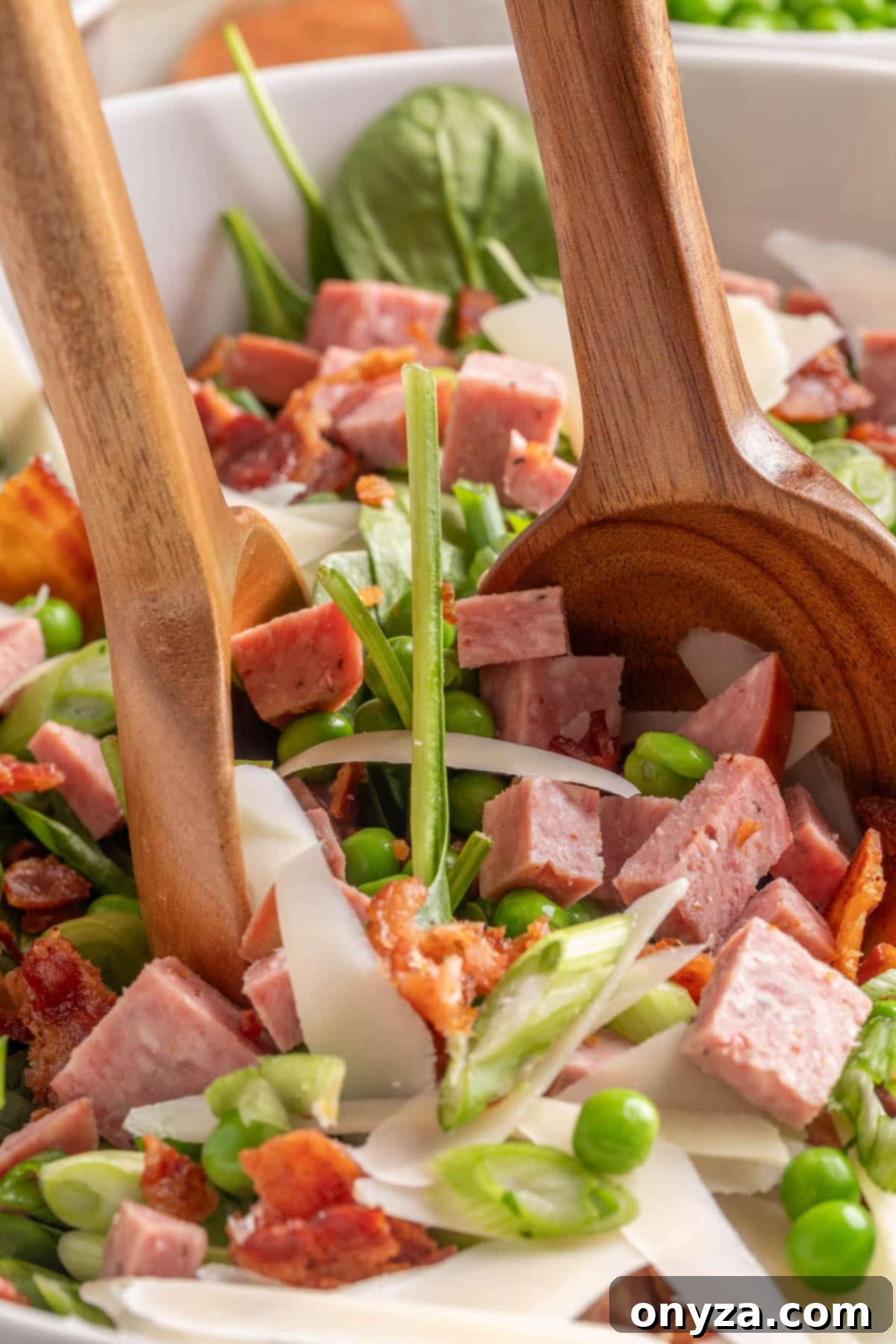 Savory Spinach & Pea Salad with Crispy Bacon and Sausage 9 Tossing a spinach and pea salad with cubed summer sausage and bacon in a white bowl with two wooden spoons, showcasing the fresh ingredients.