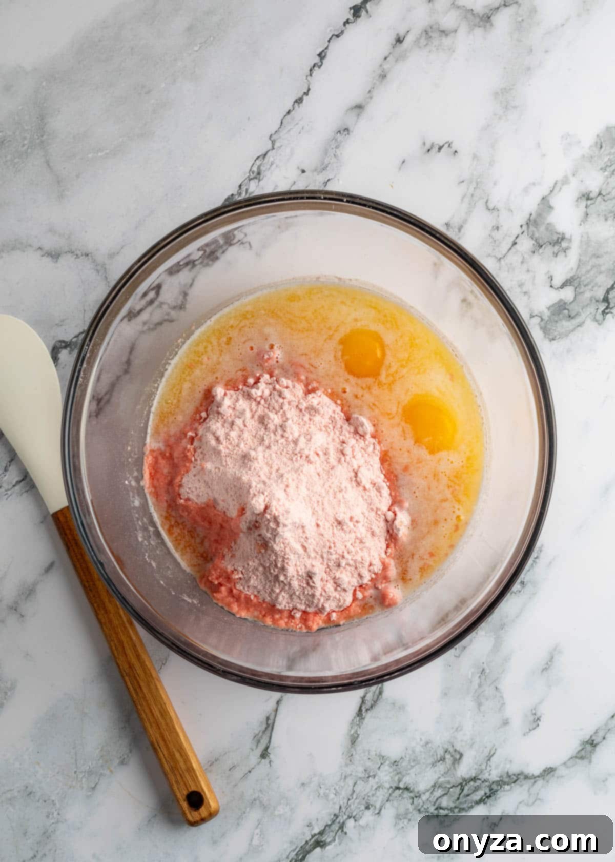 Overhead view of strawberry cake mix, melted butter, and eggs in a glass bowl on a marble board, ready to be mixed for the base layer.