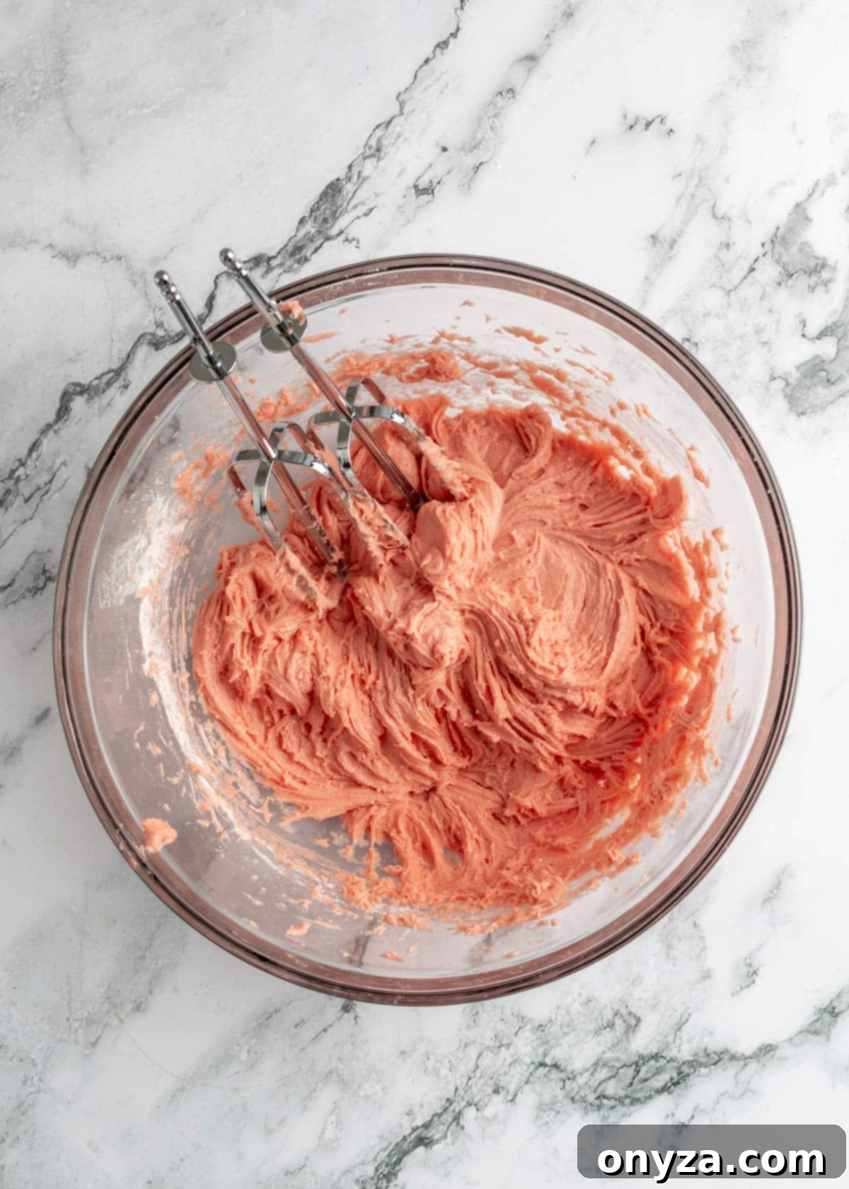 Mixed strawberry chess bar dough in a glass bowl on a white marble board, with stainless steel hand mixer beaters visible, showing the thick, smooth consistency.