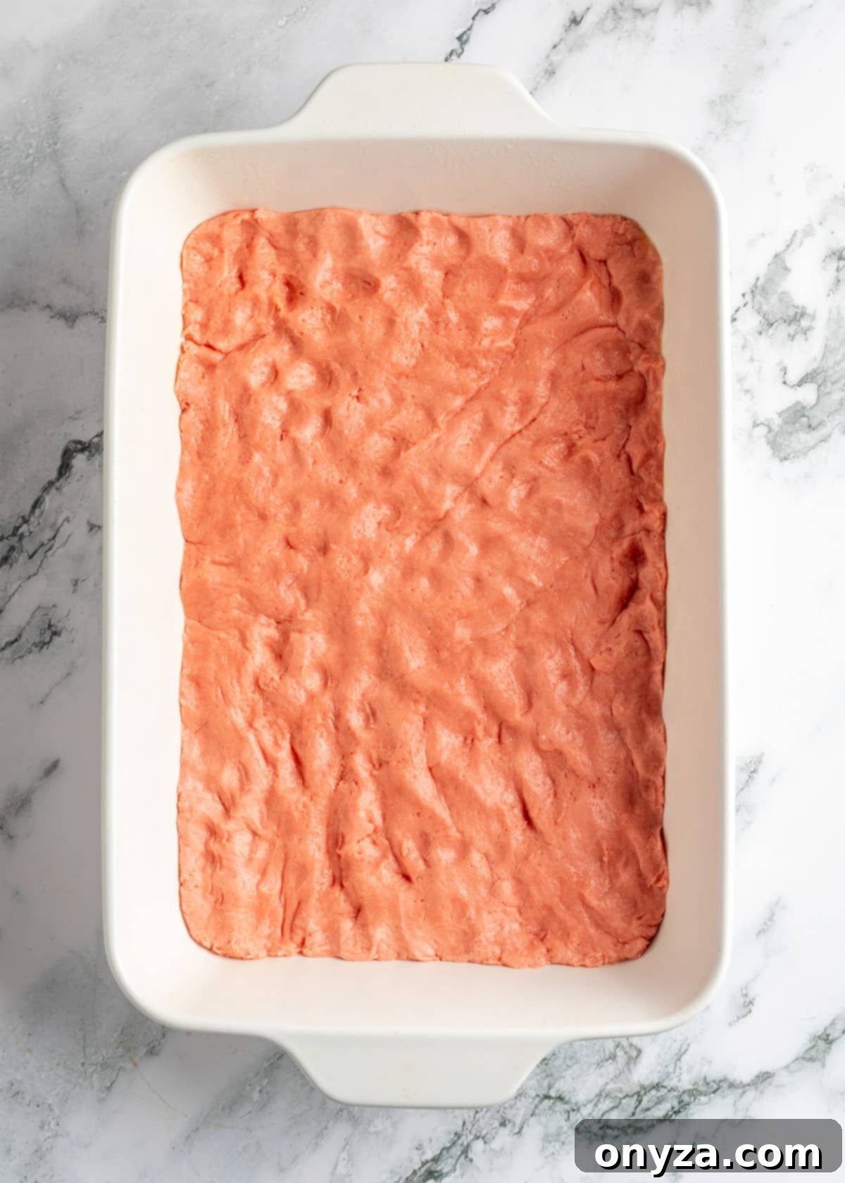 Strawberry chess bar dough pressed evenly into a white baking dish on a white marble board, forming the base layer.
