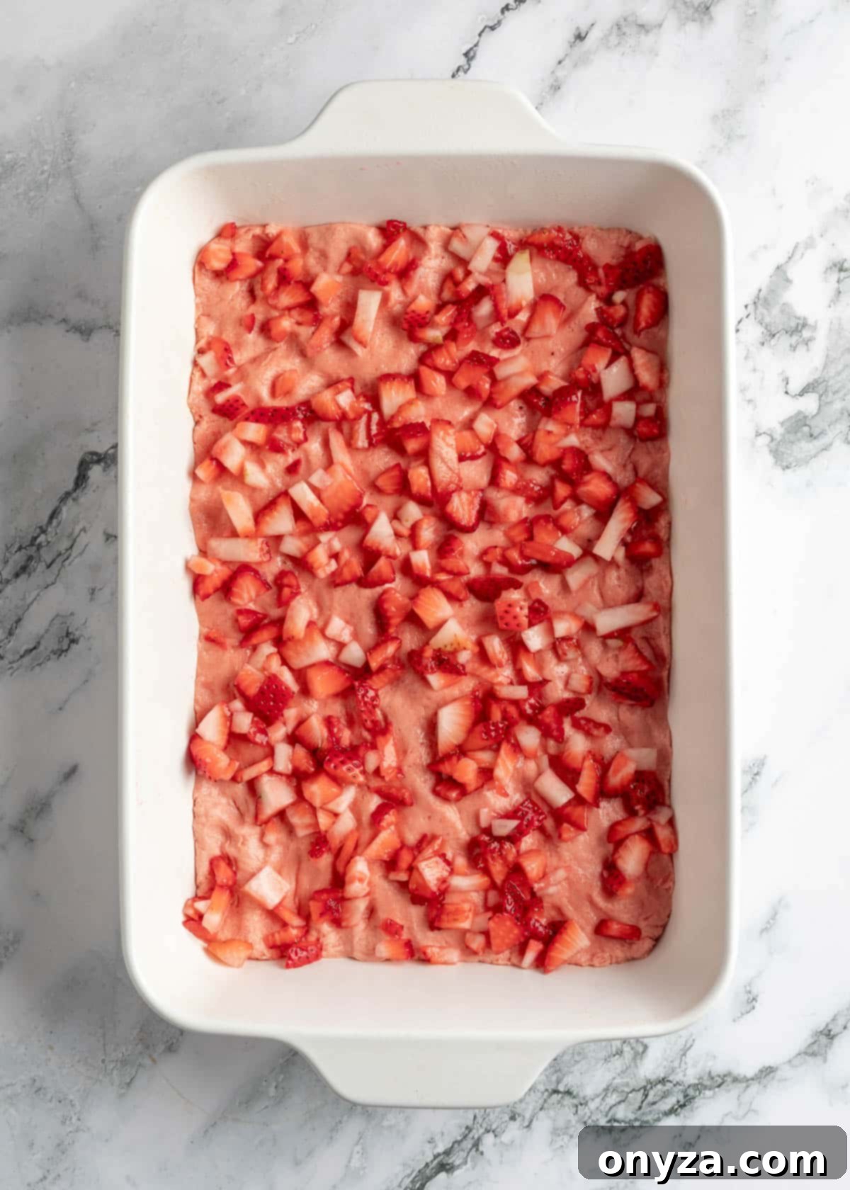 Diced strawberries evenly sprinkled over the strawberry chess square dough in a white baking dish on a white marble board, ready for the next layer.