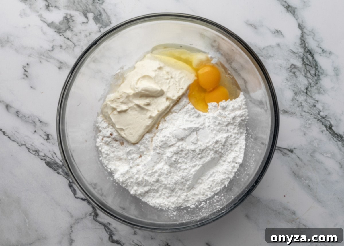 Overhead shot of cream cheese, powdered sugar, vanilla, almond extract, and eggs in a glass bowl on a white marble surface, ready for mixing to create the topping.