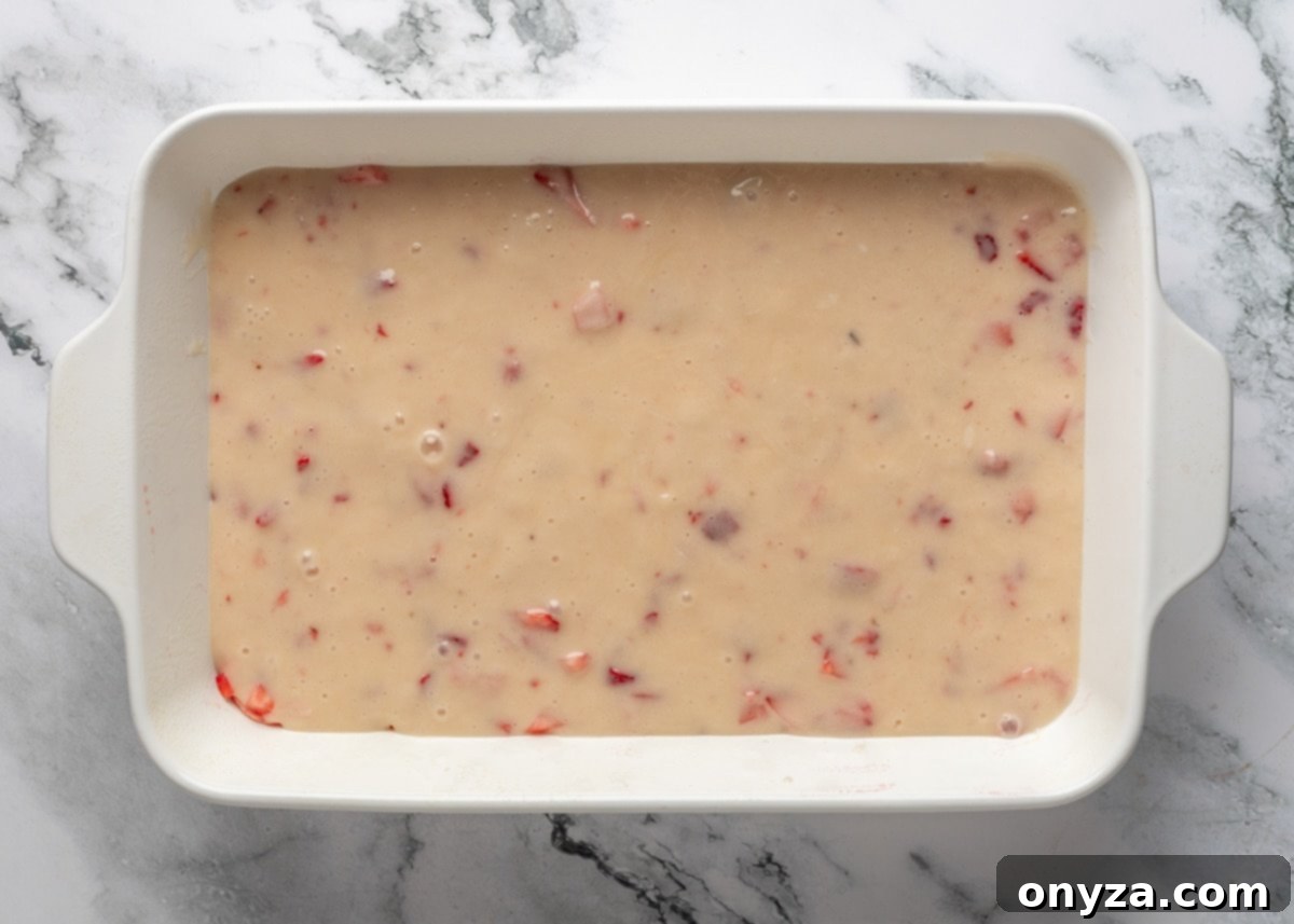 Strawberry cream cheese topping being poured over the unbaked chess squares base in a white baking dish on a white marble board, showing its smooth texture.