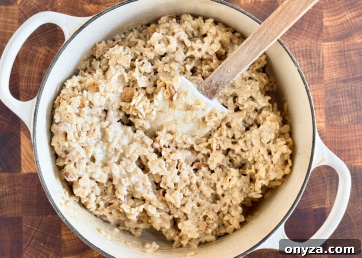 cereals mixed into apple spiced melted marshmallows in a white enameled cast iron pot with a wood handled white spatula