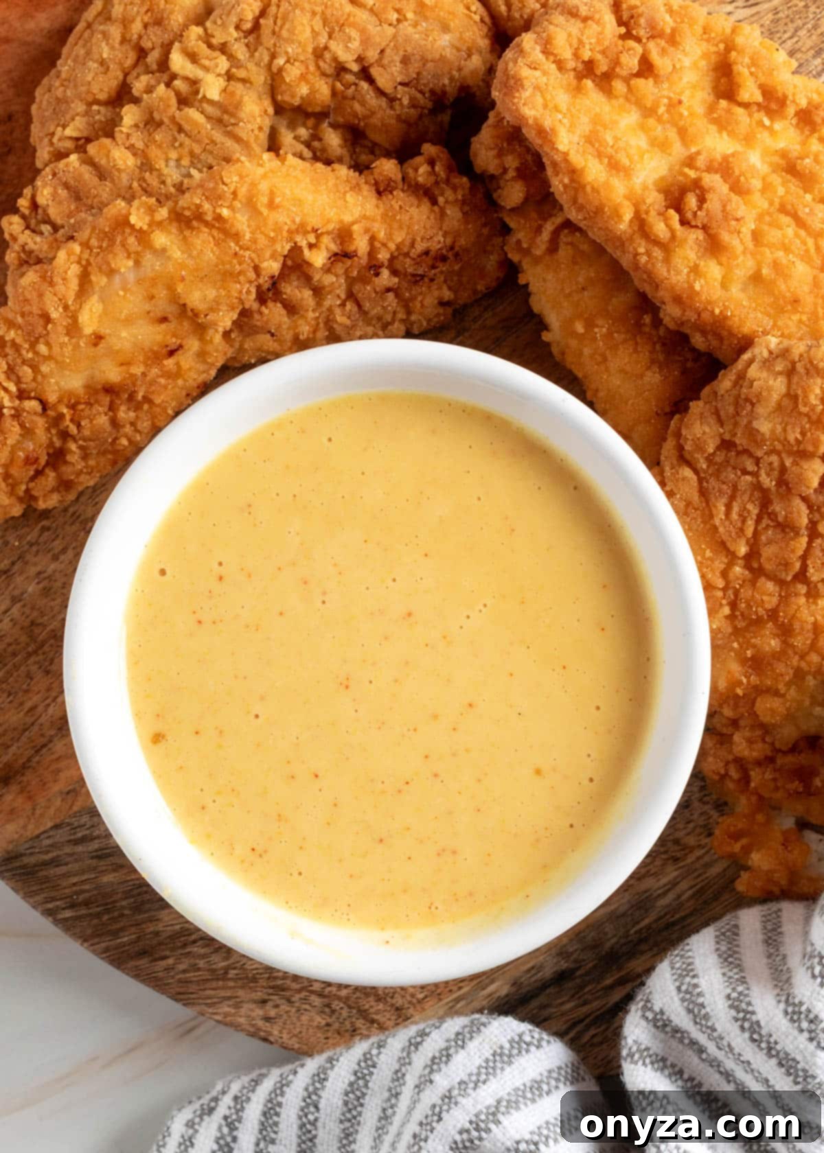 A pristine white bowl filled with glossy, golden homemade honey mustard dipping sauce is elegantly centered on a rustic round wooden board. The bowl is artfully encircled by an inviting arrangement of perfectly crispy, golden-brown fried chicken tenders, creating a tantalizing visual. A neatly folded gray-and-white striped kitchen towel subtly peeks into the frame from the corner, completing a picture of culinary comfort and impeccable style, ready for enjoyment.