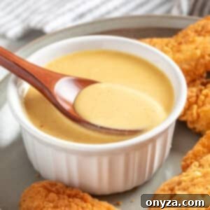 A white ramekin filled with creamy honey mustard dipping sauce on a gray plate, surrounded by crispy fried chicken tenders. A wooden spoon rests in the ramekin, lifting a spoonful of the smooth, golden-yellow sauce. A striped kitchen towel is softly blurred in the background.