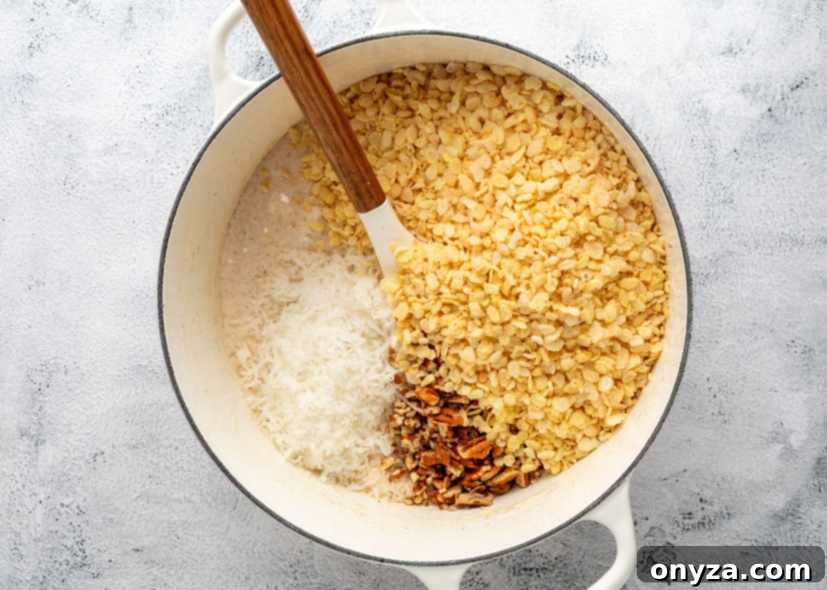Rice Krispies cereal, shredded coconut, and toasted pecans added to melted marshmallows in a white enameled cast iron pot with a wood handled spatula