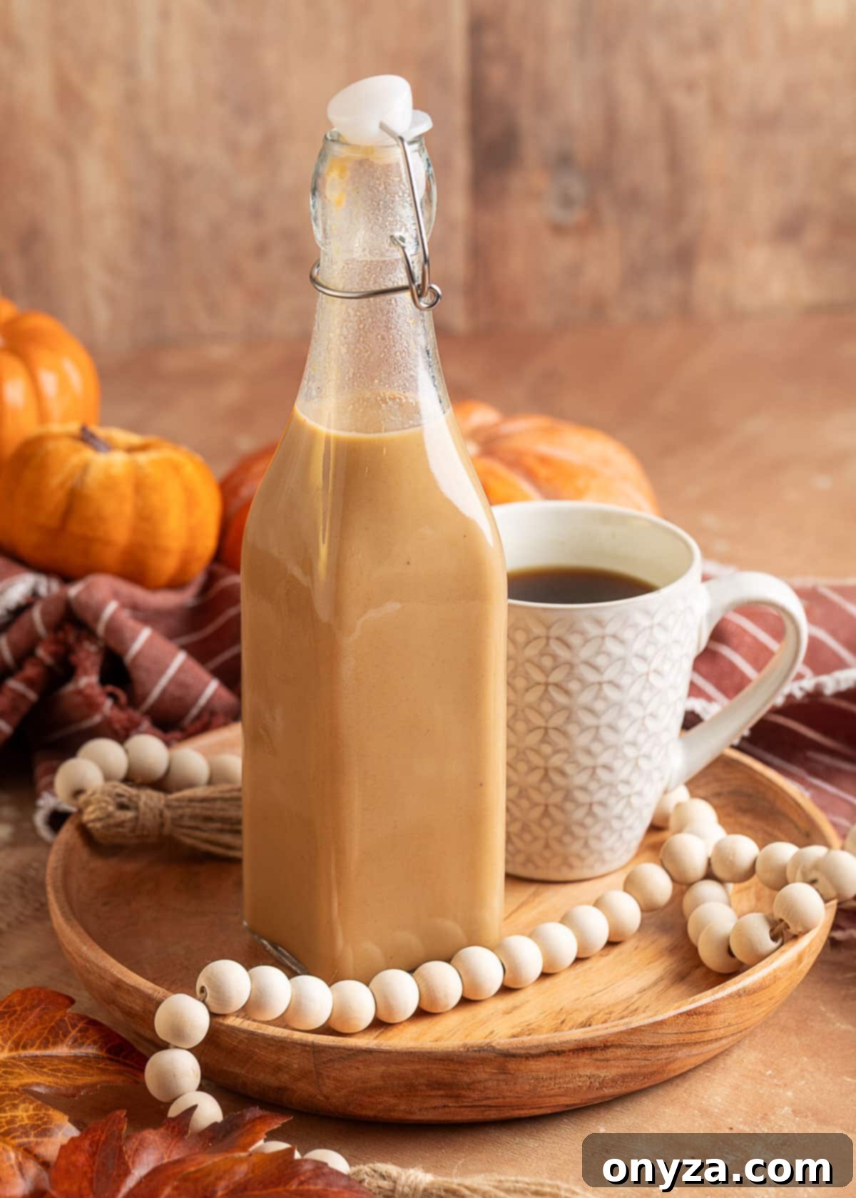 Homemade Pumpkin Spice Coffee Syrup 2 Glass swing-top bottle filled with creamy pumpkin spice coffee syrup on a wooden tray, next to a patterned white mug of black coffee. Decorative pumpkins, a striped napkin, and a wood bead garland add a fall-inspired touch.