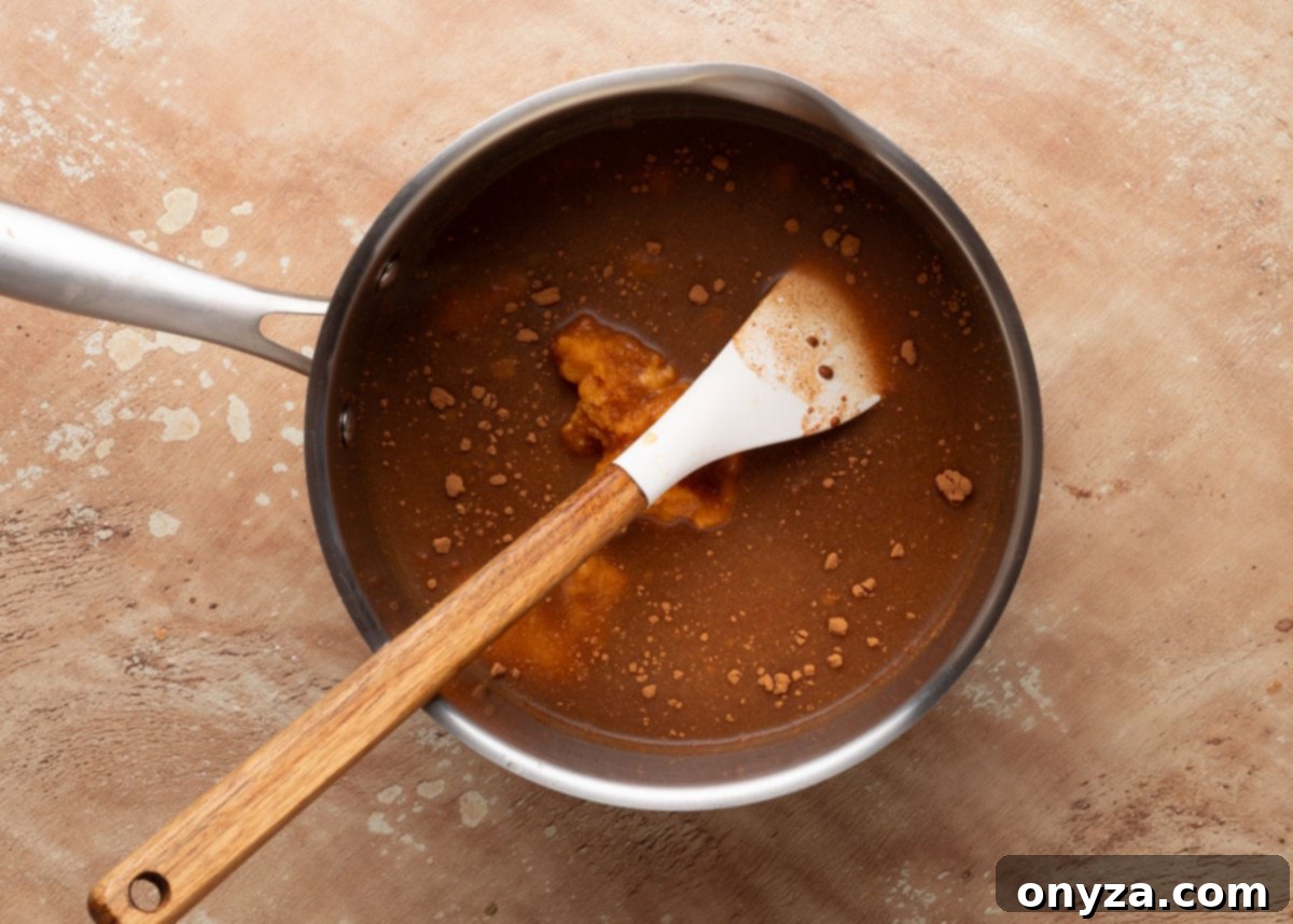 Homemade Pumpkin Spice Coffee Syrup 4 Saucepan filled with unmixed ingredients for pumpkin spice coffee syrup, including visible pumpkin purée and spices, with a white spatula resting inside the pan on a tan surface.