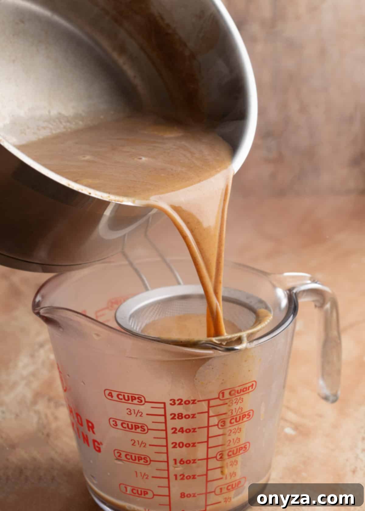 Homemade Pumpkin Spice Coffee Syrup 5 Warm pumpkin spice coffee syrup being poured from a saucepan through a fine mesh strainer into a glass measuring cup. Steam rises from the syrup against a brown background.