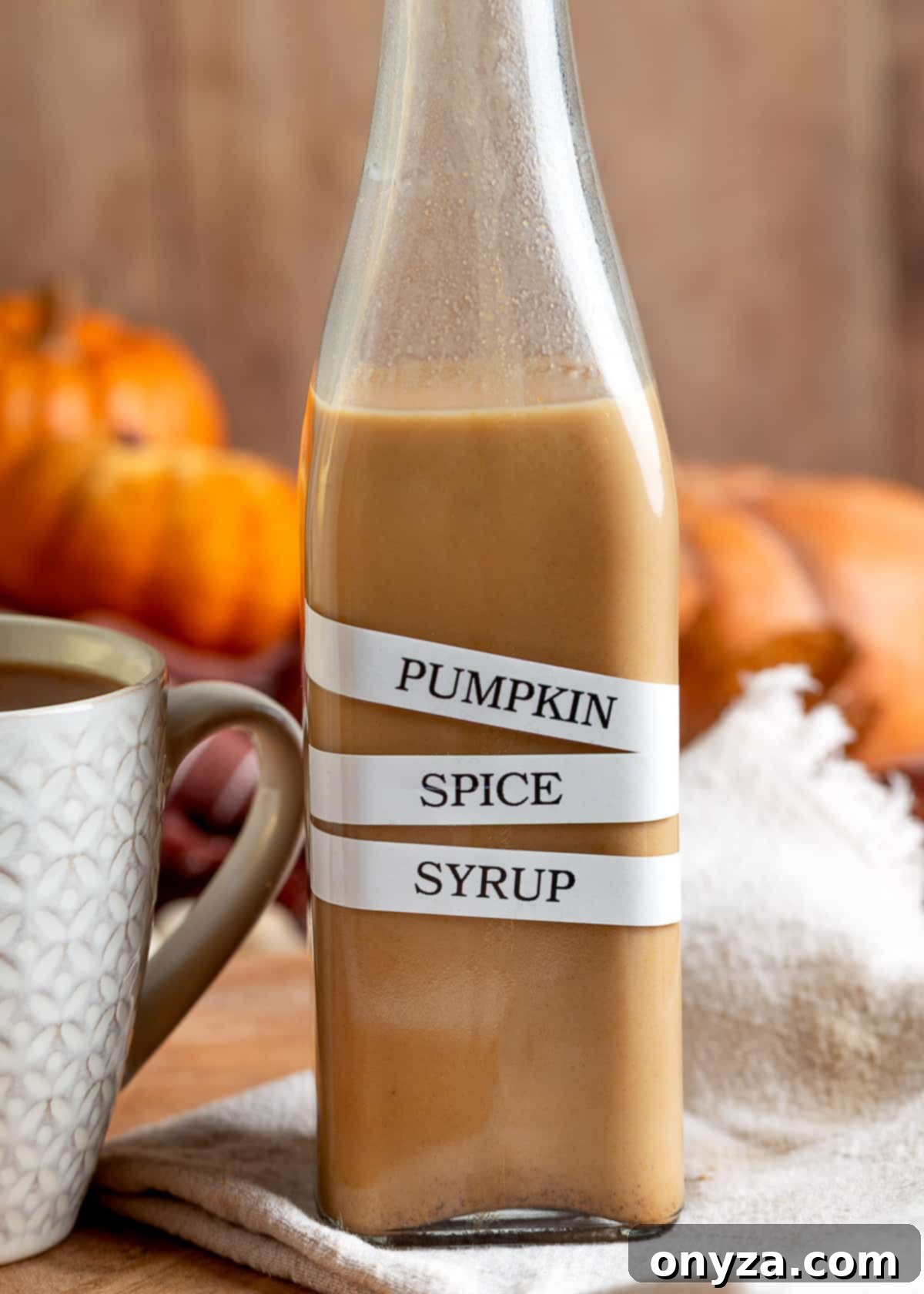 Homemade Pumpkin Spice Coffee Syrup 6 A close-up of a bottle labeled “Pumpkin Spice Syrup” in bold, staggered black lettering. The golden syrup fills the bottle with a warm hue, and a ceramic coffee mug and small pumpkins are softly visible behind it.
