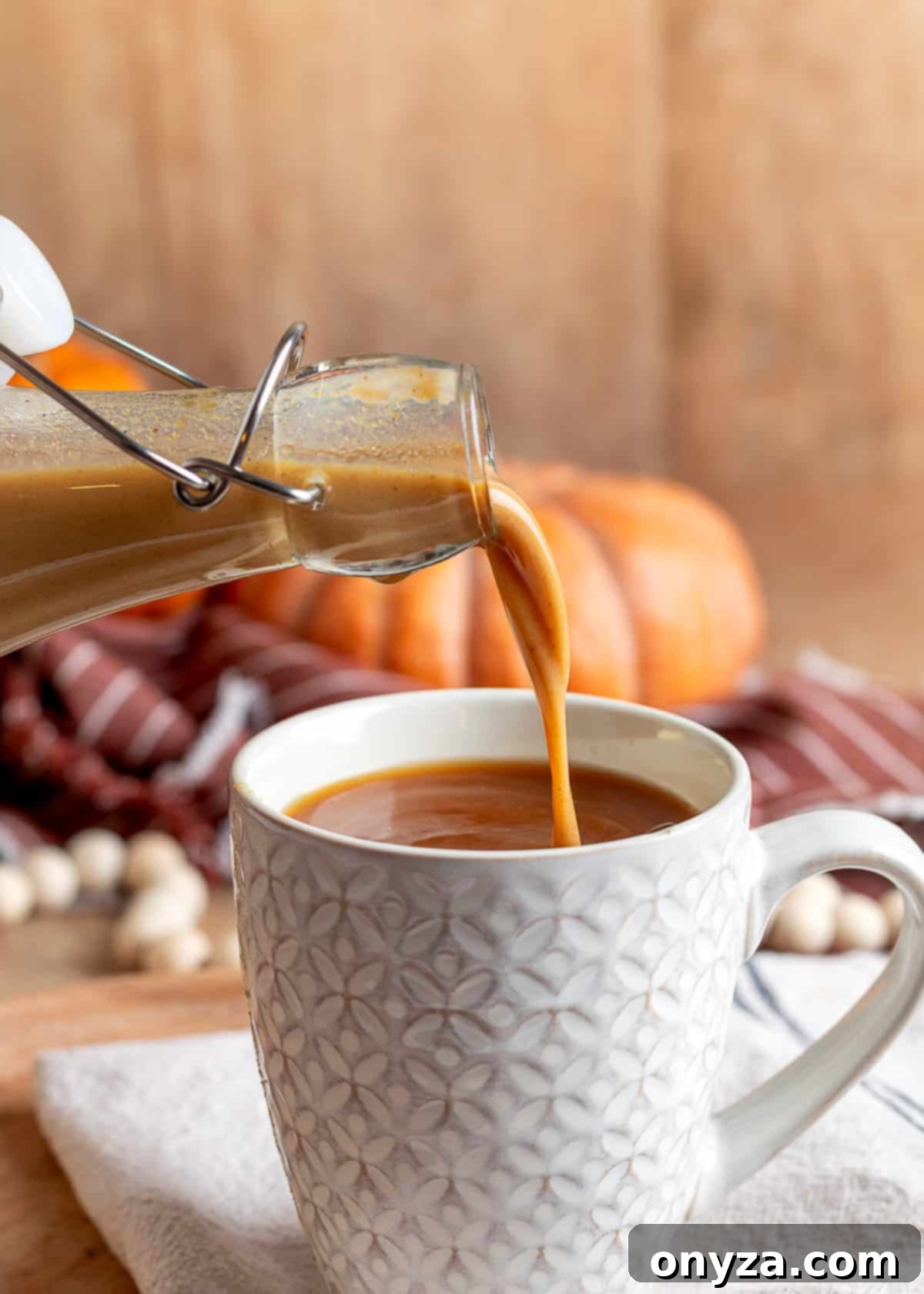 Homemade Pumpkin Spice Coffee Syrup 7 Pumpkin spice syrup being poured from a glass bottle into a mug of coffee. A brown striped napkin, white wood-bead garland, and small pumpkins are softly blurred in the fall-themed background.