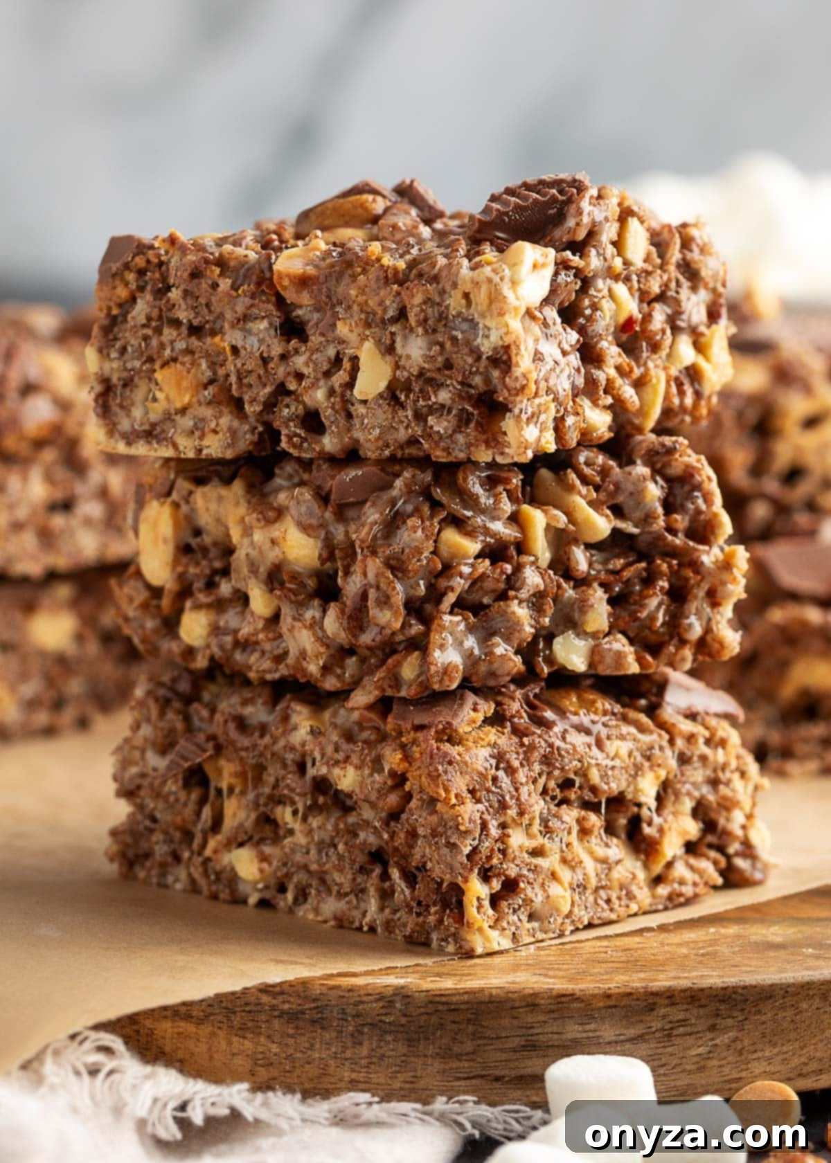 Stack of three peanut butter cup rice krispie treats on a wooden board lined with parchment paper. The bars are thick and chewy, filled with chocolate puffed rice cereal, chopped peanuts, and bits of peanut butter cups. A few mini marshmallows and peanut butter chips are scattered at the bottom of the frame.
