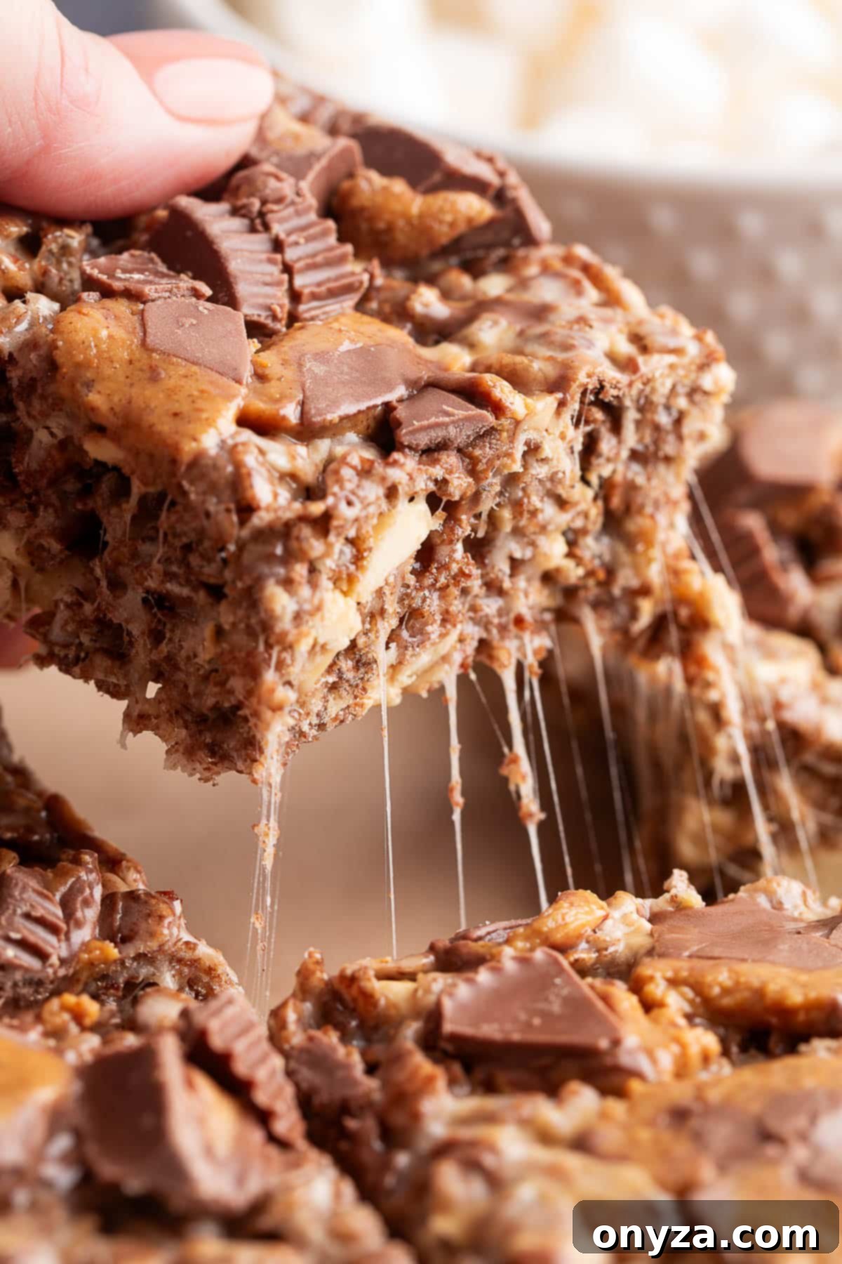 Close-up of a hand lifting a gooey peanut butter cup rice krispie treat from a batch resting on parchment paper. The bar is studded with chopped peanut butter cups and peanuts, with visible strands of melted marshmallow stretching as the treat is pulled away.