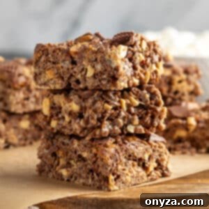 Stack of three peanut butter cup rice krispie treats on a wooden board lined with parchment paper. The bars are thick and chewy, filled with chocolate puffed rice cereal, chopped peanuts, and bits of peanut butter cups. A few mini marshmallows and peanut butter chips are scattered at the bottom of the frame.
