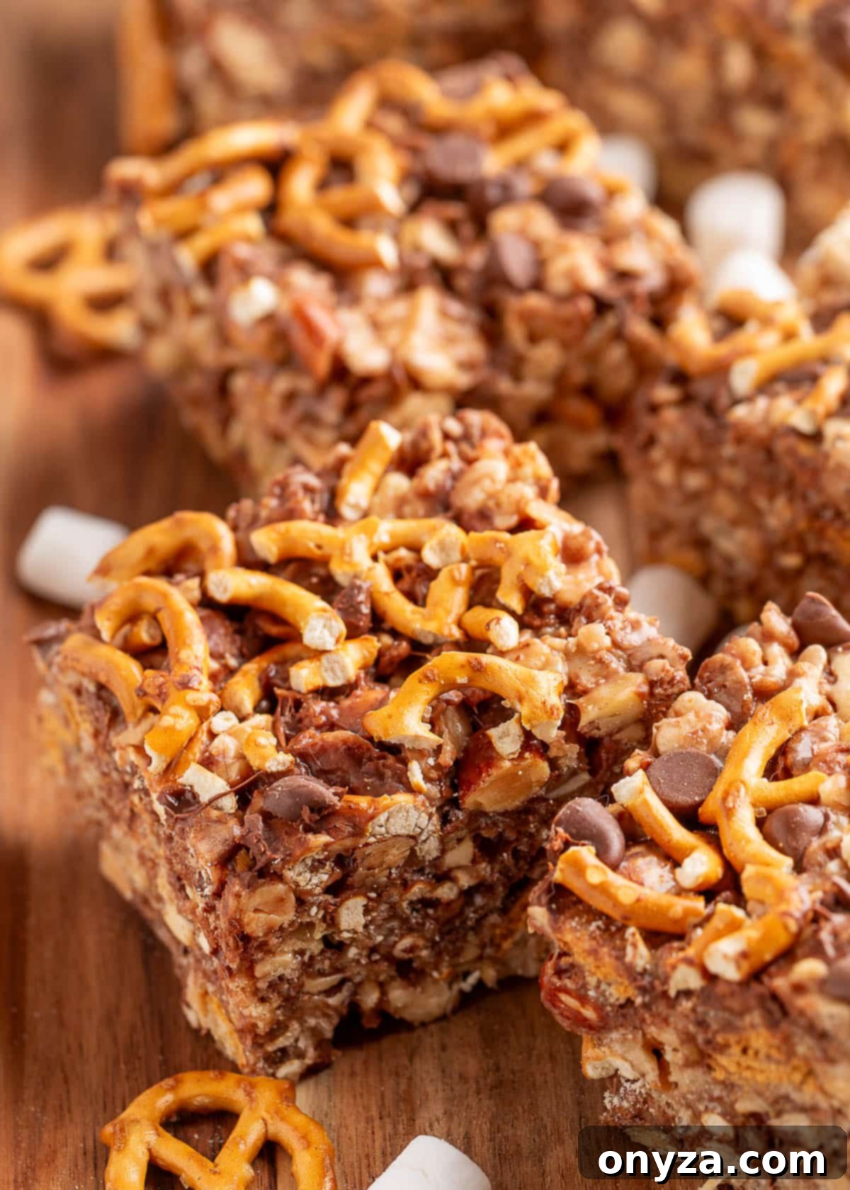 Close-up of loaded rice krispie treats cut into squares on a wooden board. The bars are packed with pretzel twists, chocolate chips, nuts, and marshmallows, creating a sweet and salty texture. Pieces of broken pretzels and mini marshmallows are scattered around the board.