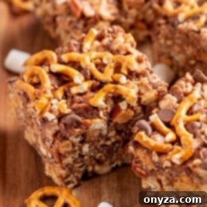 Close-up of kitchen sink rice krispie treats cut into squares on a wooden board. The bars are packed with pretzel twists, chocolate chips, nuts, and marshmallows, creating a sweet and salty texture. Pieces of broken pretzels and mini marshmallows are scattered around the board.