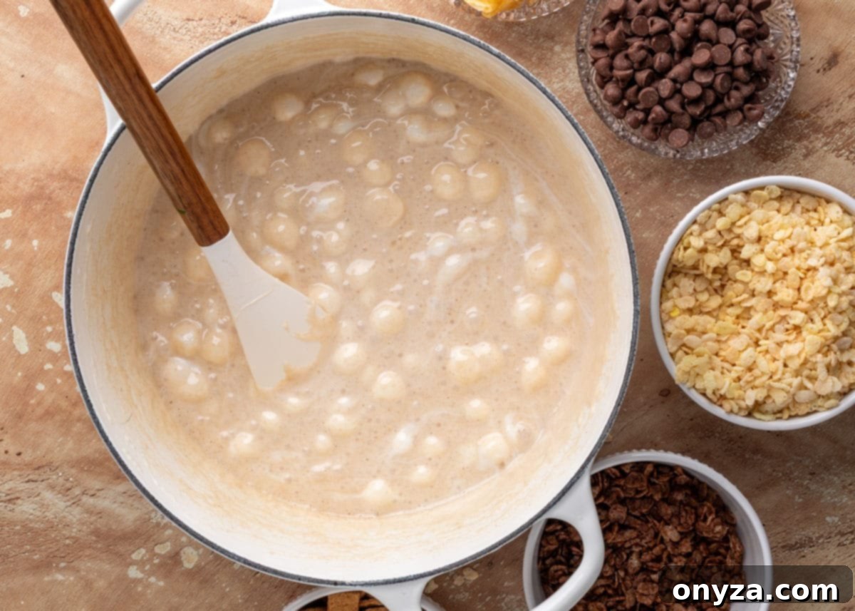 Melted marshmallows in a white Dutch oven stirred with a spatula, creating a gooey base. Bowls of cereals, chips, and chocolate chips sit nearby, ready to be mixed in.