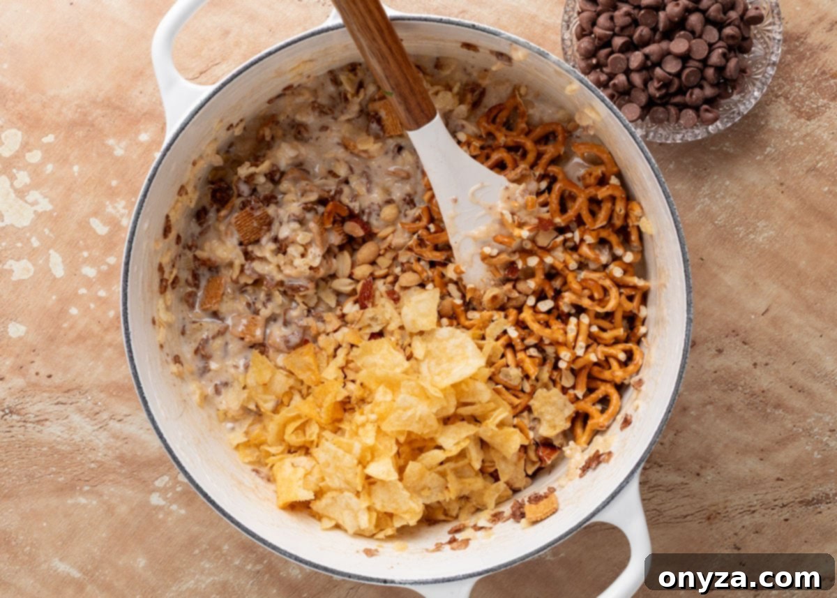 Crushed potato chips and mini pretzels are added to the partially mixed cereal and marshmallow base in the pot. The chocolate chips remain in a bowl to the side.