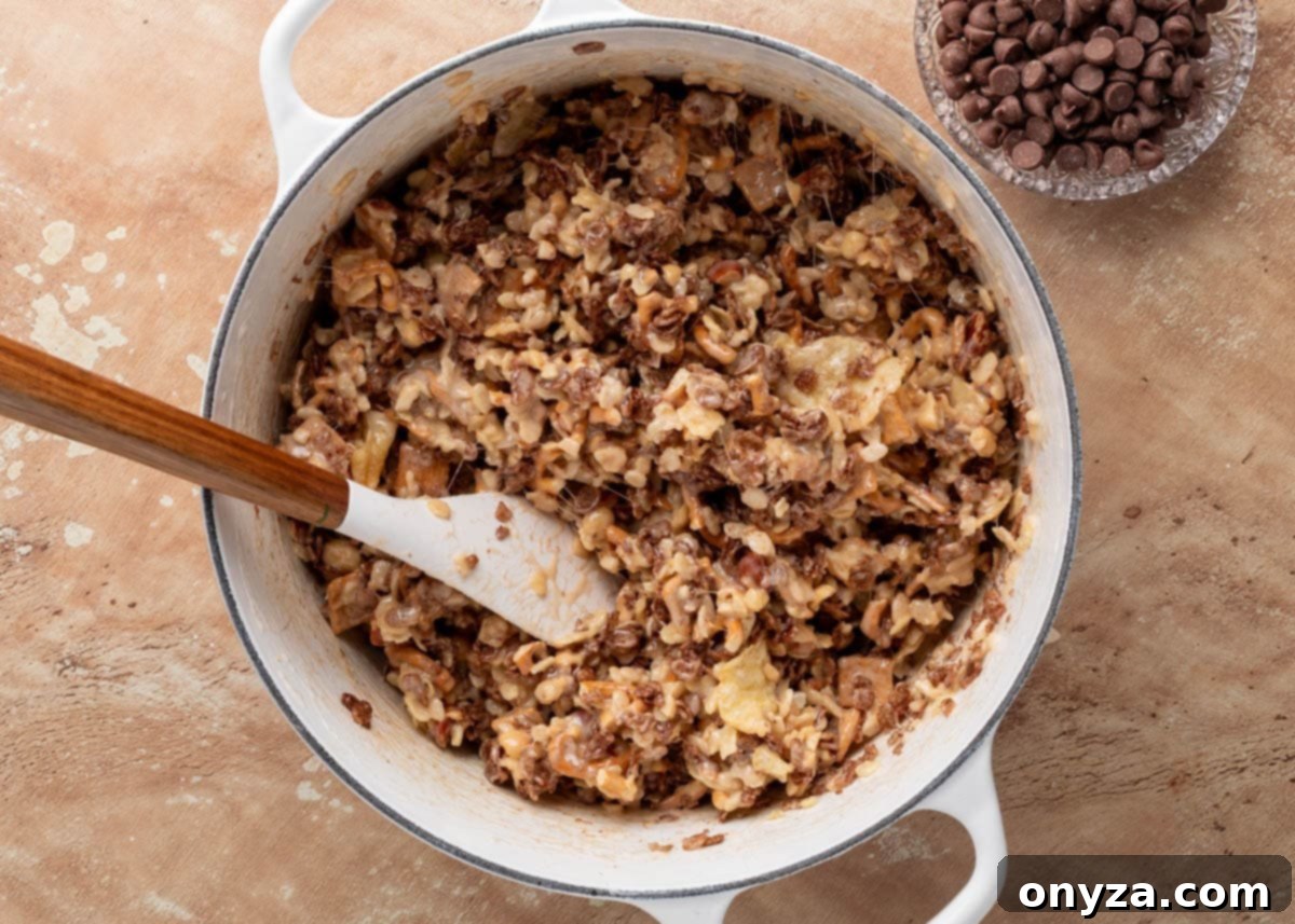 The fully mixed base for loaded rice krispie treats in a Dutch oven, combining cereals, chips, pretzels, and gooey marshmallow. A spatula rests in the mixture, and the chocolate chips are still set aside, ready to be stirred in last.