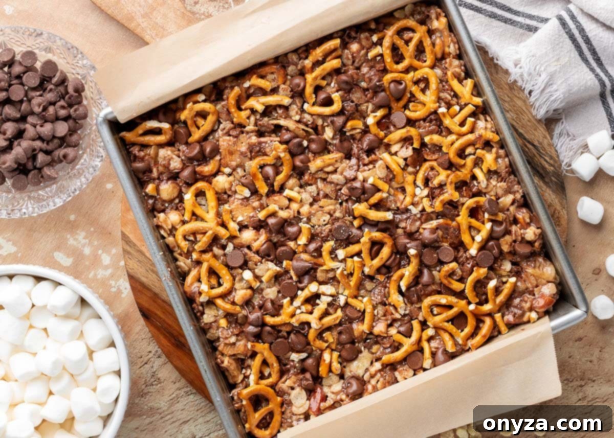 Overhead view of uncut loaded rice krispie treats in a parchment-lined metal pan. The surface is topped with mini pretzels and semisweet chocolate chips, pressed into a gooey mixture of cereal, marshmallows, and crushed snacks. Bowls of chocolate chips and marshmallows are visible nearby on the table.