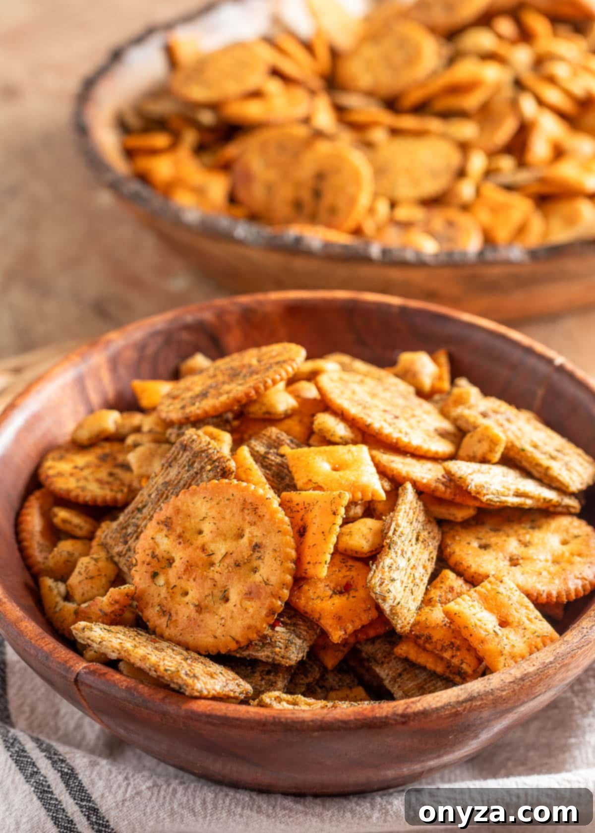 Zesty Dill Ranch Crackers 2 Close-up of a wooden bowl filled with homemade dill ranch crackers, featuring seasoned Ritz-style crackers, cheese crackers, and woven wheat squares, with a second bowl of the mix blurred in the background.