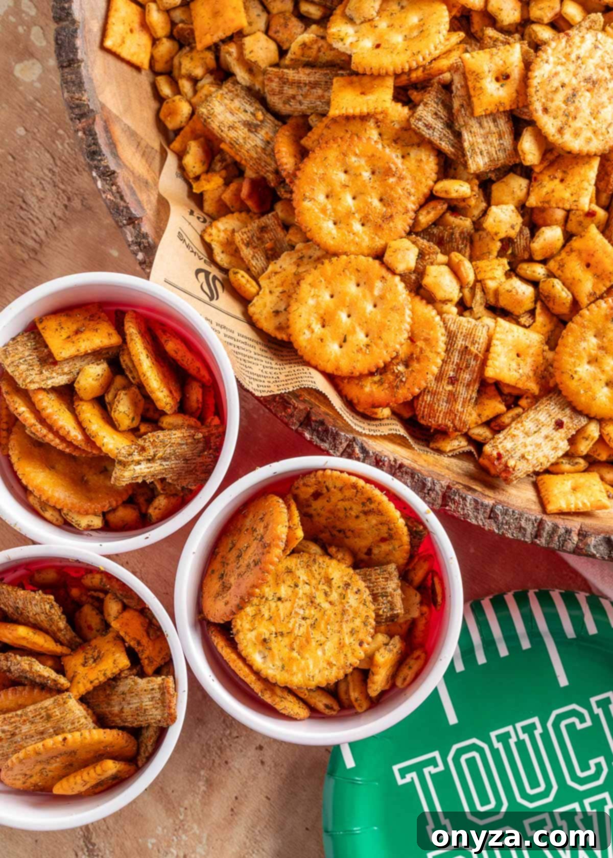 Zesty Dill Ranch Crackers 7 Overhead view of a rustic wood bowl and several white party cups filled with dill ranch snack mix, including cheese crackers, wheat crackers, butter crackers, and oyster crackers. A green football-themed paper plate labeled “Touchdown” is partially visible in the corner, suggesting a game day setting.