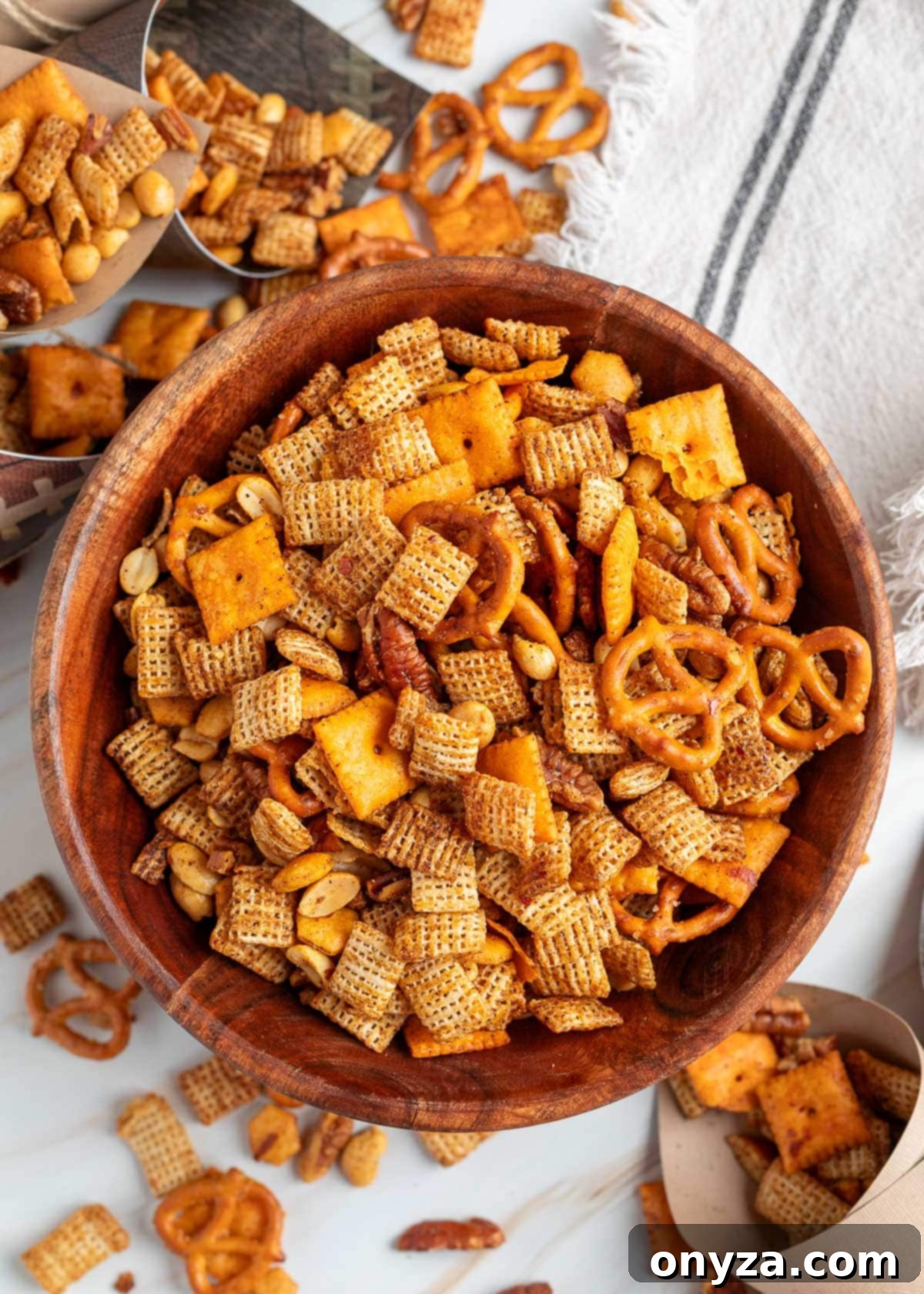 Overhead view of a large wooden bowl filled with BBQ snack mix, including pretzels, rice Chex, cheese crackers, peanuts, and pecans. The snack mix is golden brown and seasoned, with additional mix scattered on the white surface and in small paper cones in the background.
