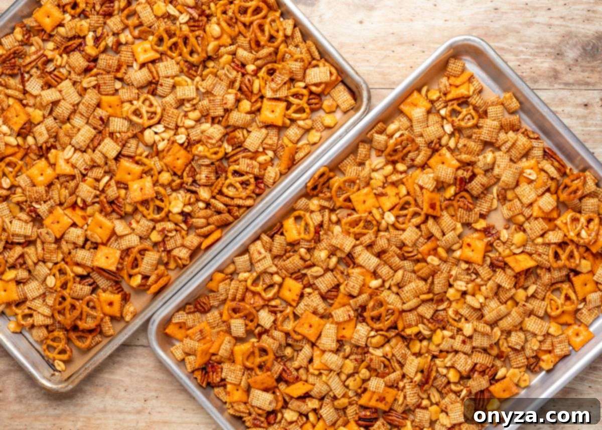 Two sheet pans filled with BBQ snack mix, including Chex cereal, Cheez-It crackers, pretzels, peanuts, and pecans, coated in a golden seasoning and arranged on parchment paper.