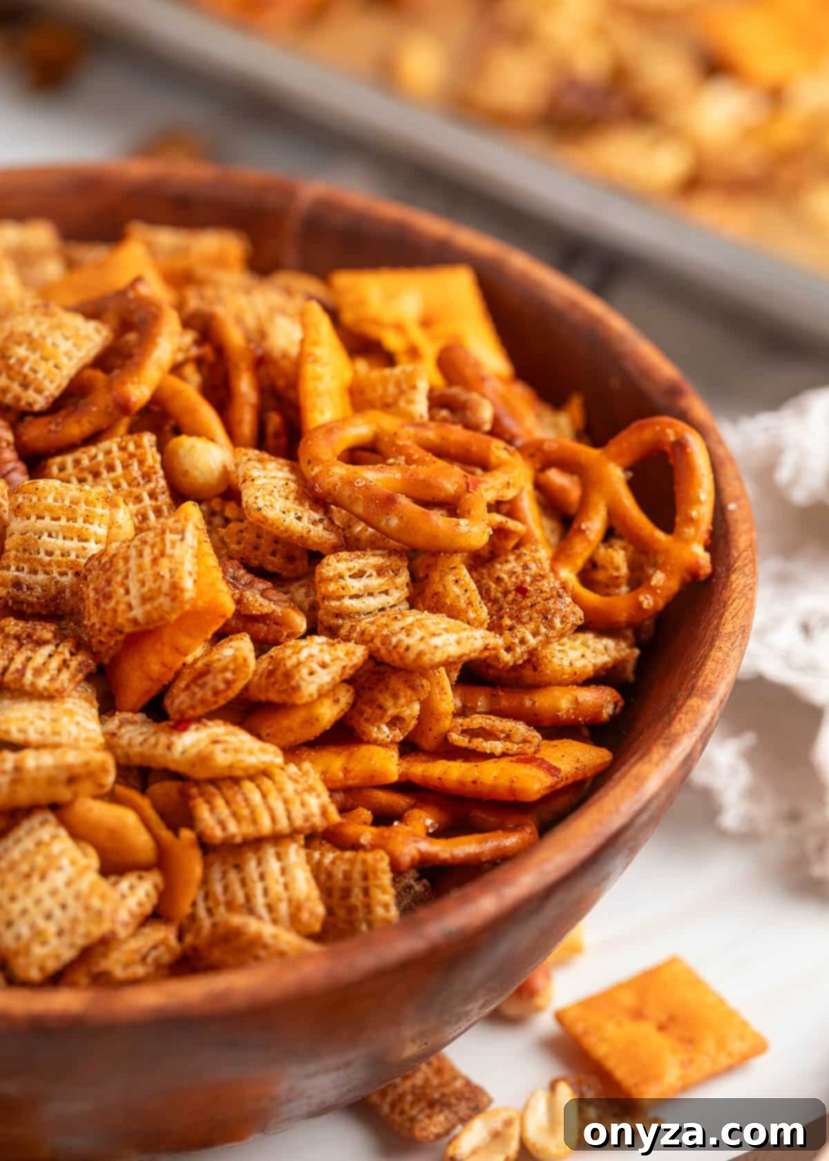 A close-up of BBQ snack mix in a rustic wooden bowl, filled with seasoned Chex cereal, mini pretzels, cheese crackers, peanuts, and other crunchy pieces. A baking sheet of the same mix is blurred in the background.