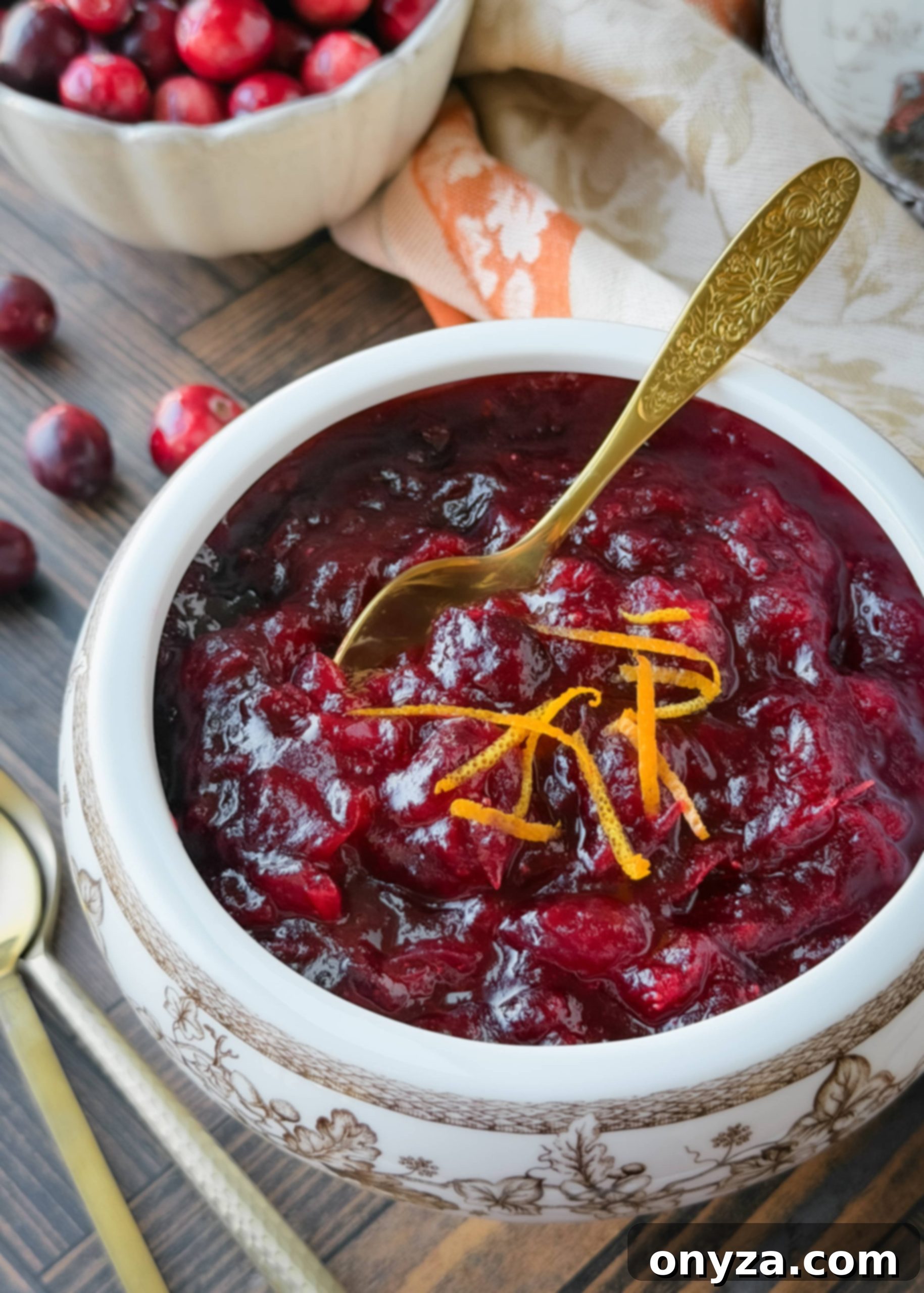 Whole berry cranberry sauce in a white and brown floral bowl, garnished with orange zest and served with a vintage gold spoon; fresh cranberries and autumn linens in the background.