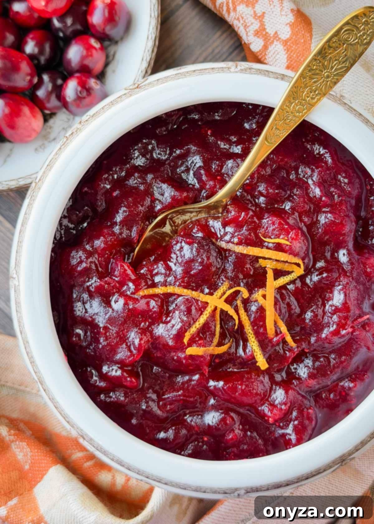Overhead view of a white bowl filled with classic whole berry cranberry sauce, garnished with thin strips of orange zest and served with a gold floral-handled spoon; fresh cranberries and an autumn-toned napkin are partially visible in the background.