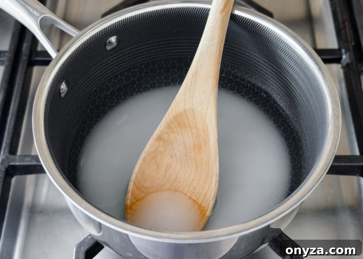 A saucepan on a stovetop with a wooden spoon stirring a cloudy mixture of sugar and water as it begins to dissolve.