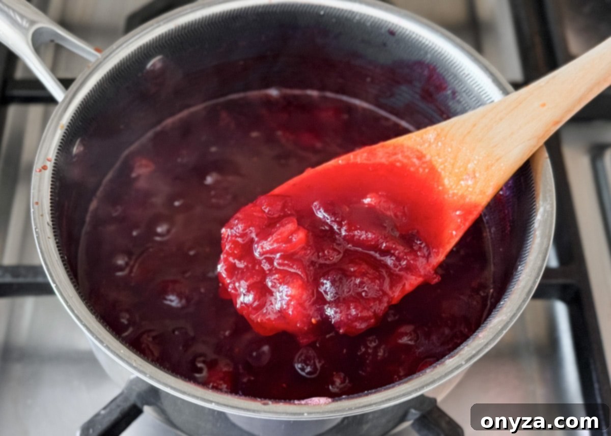 A close-up of thickened cranberry sauce in the saucepan, with a wooden spoon lifting a scoop to show texture.