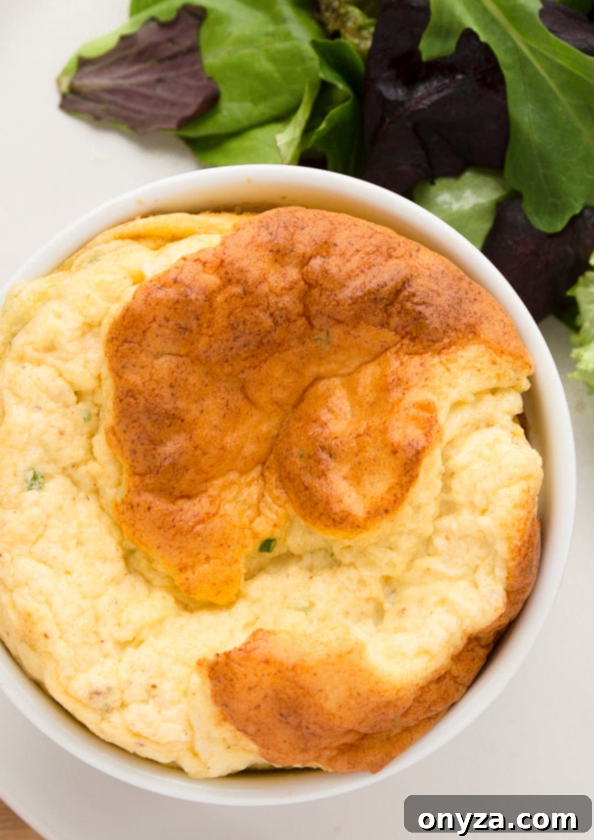 Overhead shot of a beautifully baked roasted cauliflower soufflé, served in its ramekin on a white plate with a vibrant green salad, showcasing its golden-brown top.