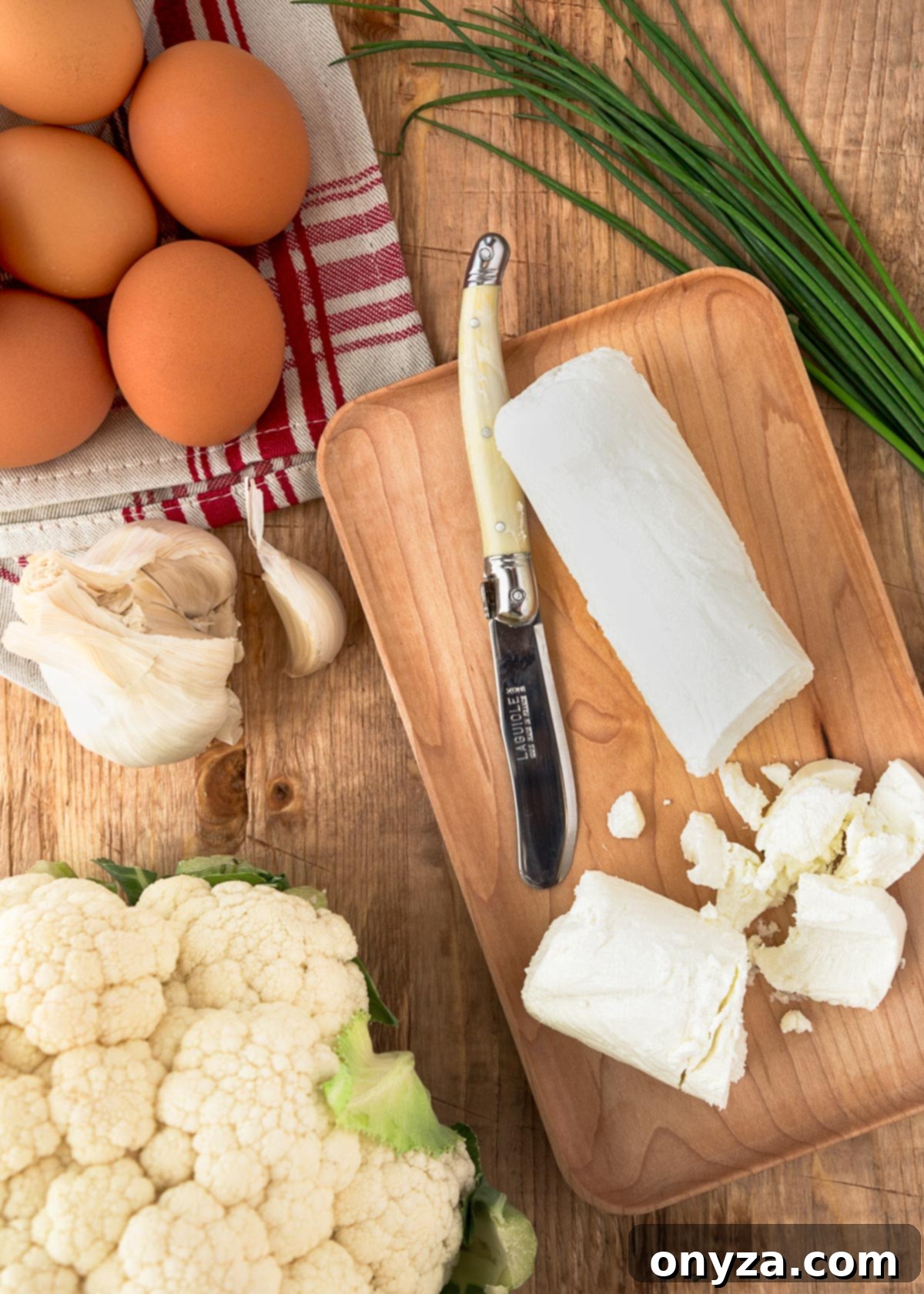 Key ingredients for cauliflower soufflés including goat cheese, brown eggs, a head of garlic, fresh chives, and a whole cauliflower head, arranged on a rustic wood board.