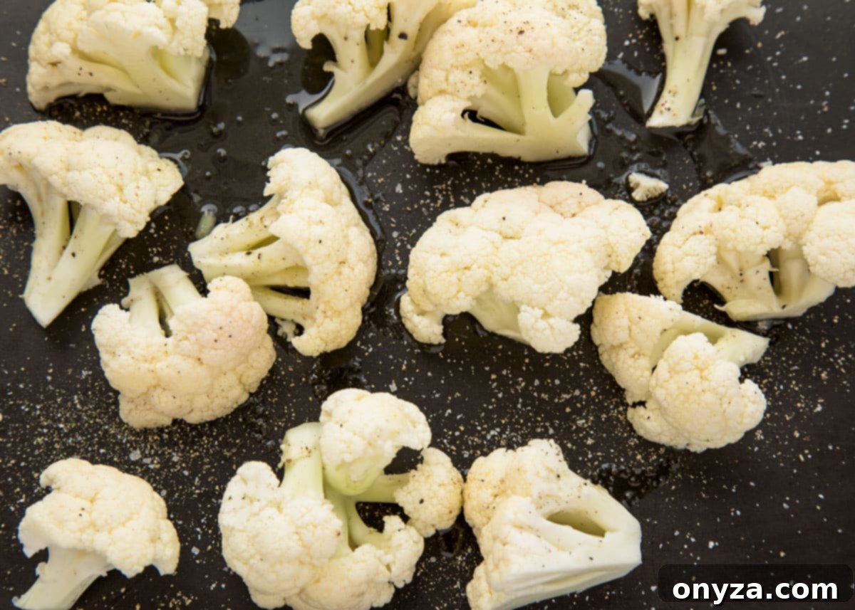 Raw cauliflower florets meticulously arranged on a black baking sheet, lightly coated with olive oil, salt, and pepper, ready for roasting.