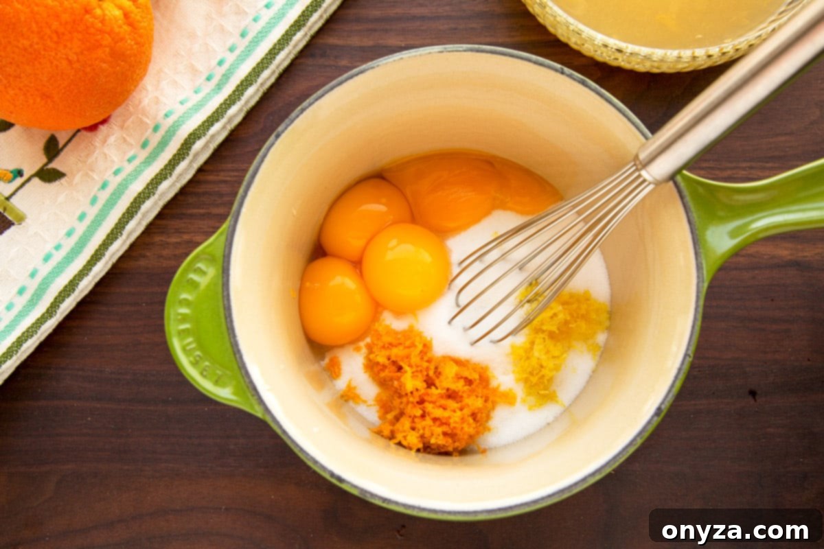Egg yolks, granulated sugar, and citrus zest combined in a green and white saucepan with a stainless steel whisk, illustrating the initial steps of making orange curd.